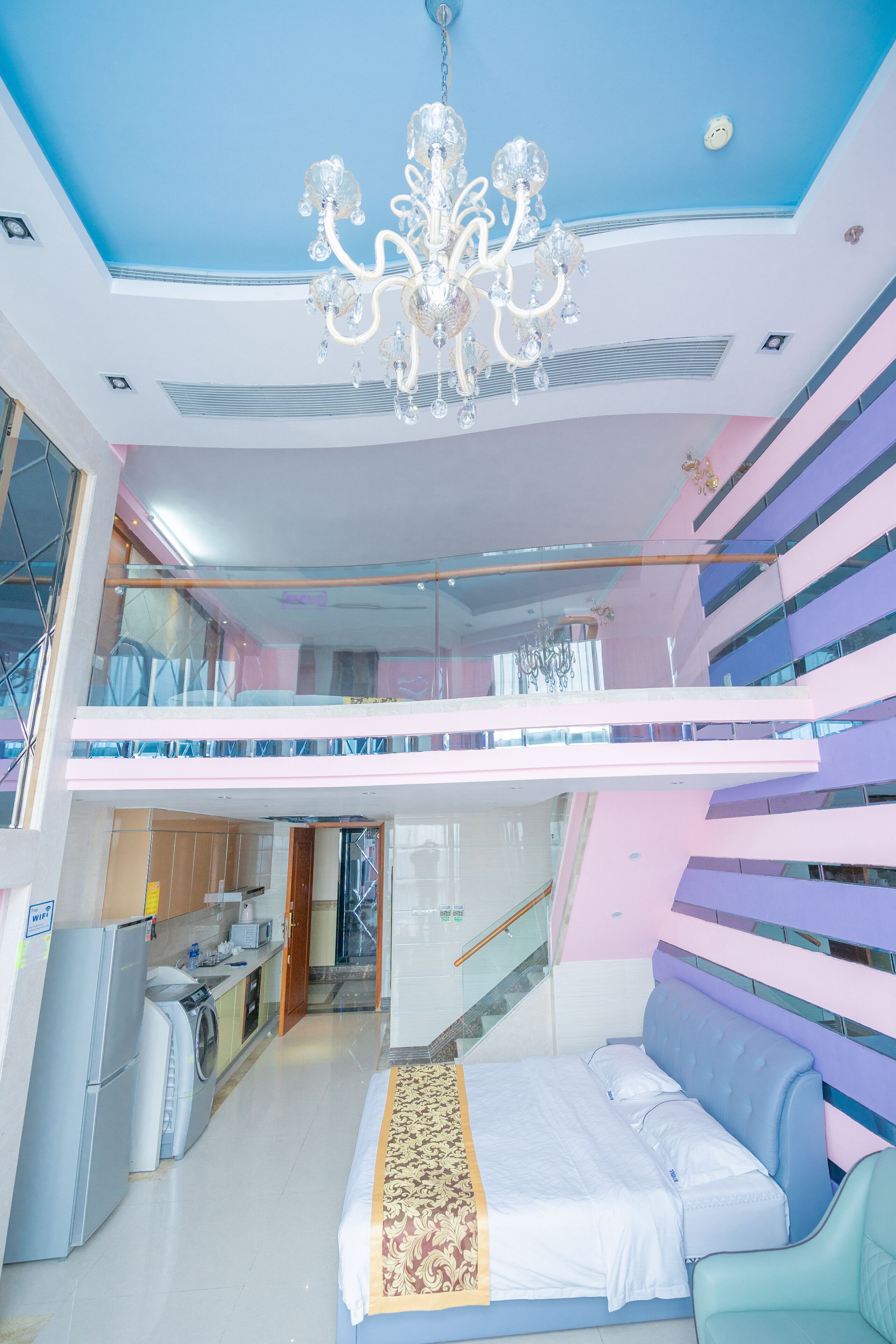 A modern hotel room with a large bed, a chandelier, a balcony, and a kitchen area.