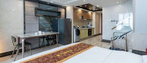 A hotel room with a bed, a desk with chairs, a washing machine, and a kitchen area.