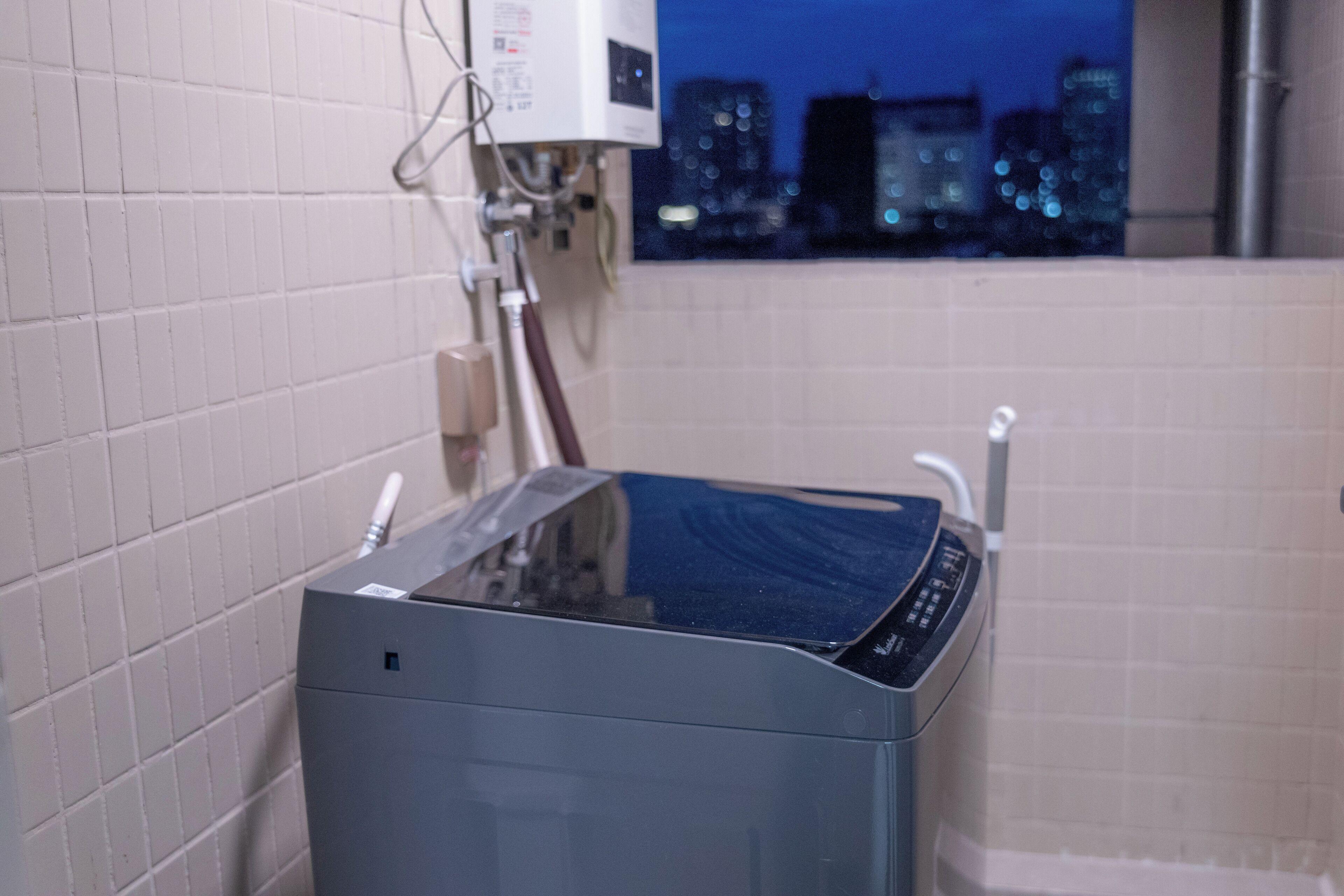 A modern bathroom with a built-in washer and dryer, white tiled walls, and a view of a cityscape at night.