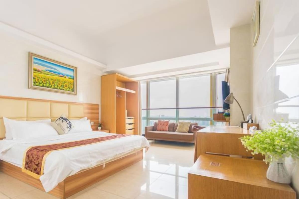 A modern hotel room with a large bed, a desk, a sofa, and a large window offering a city view.