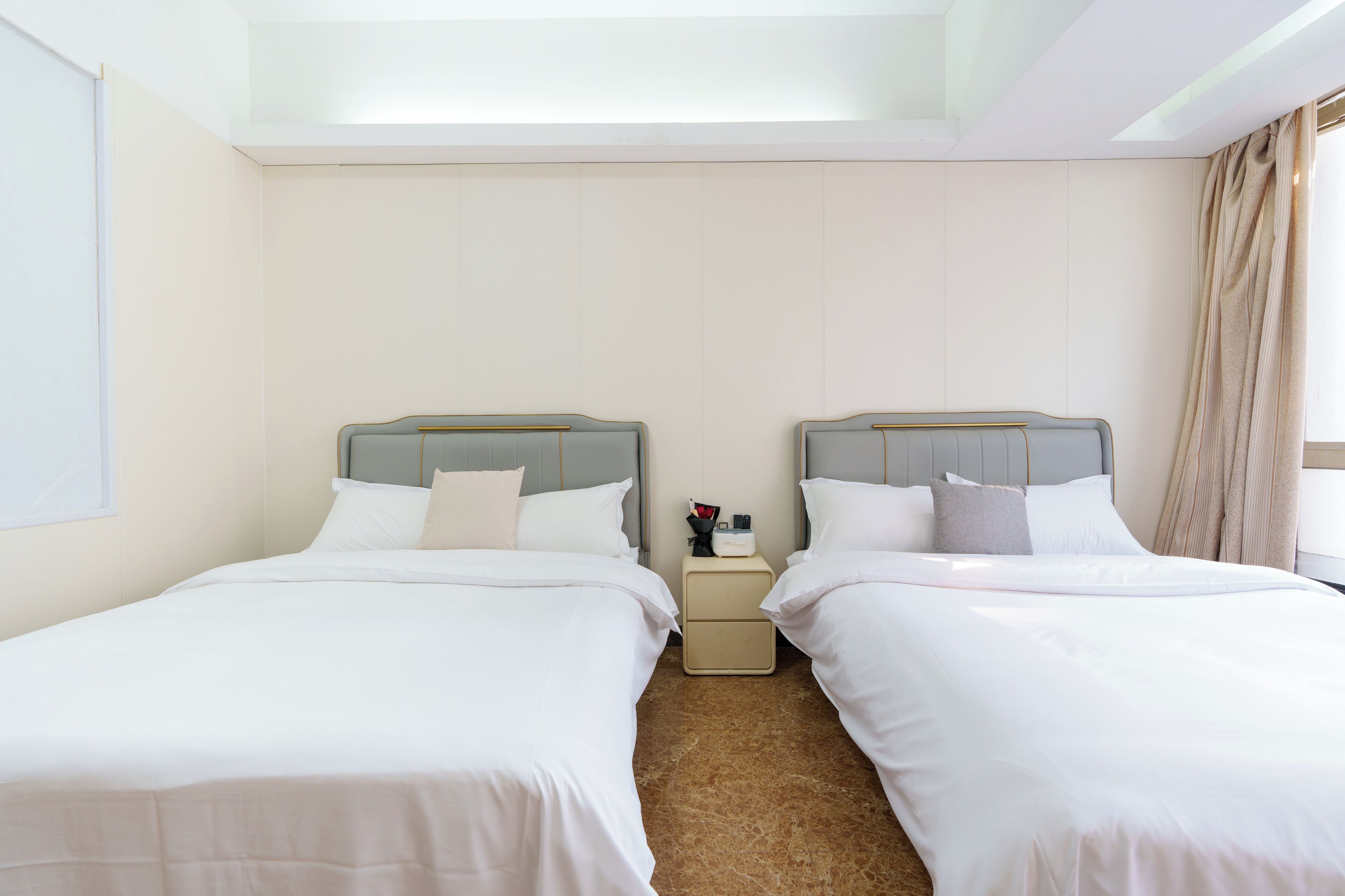 A hotel room with two beds, a nightstand, and a large window with curtains.