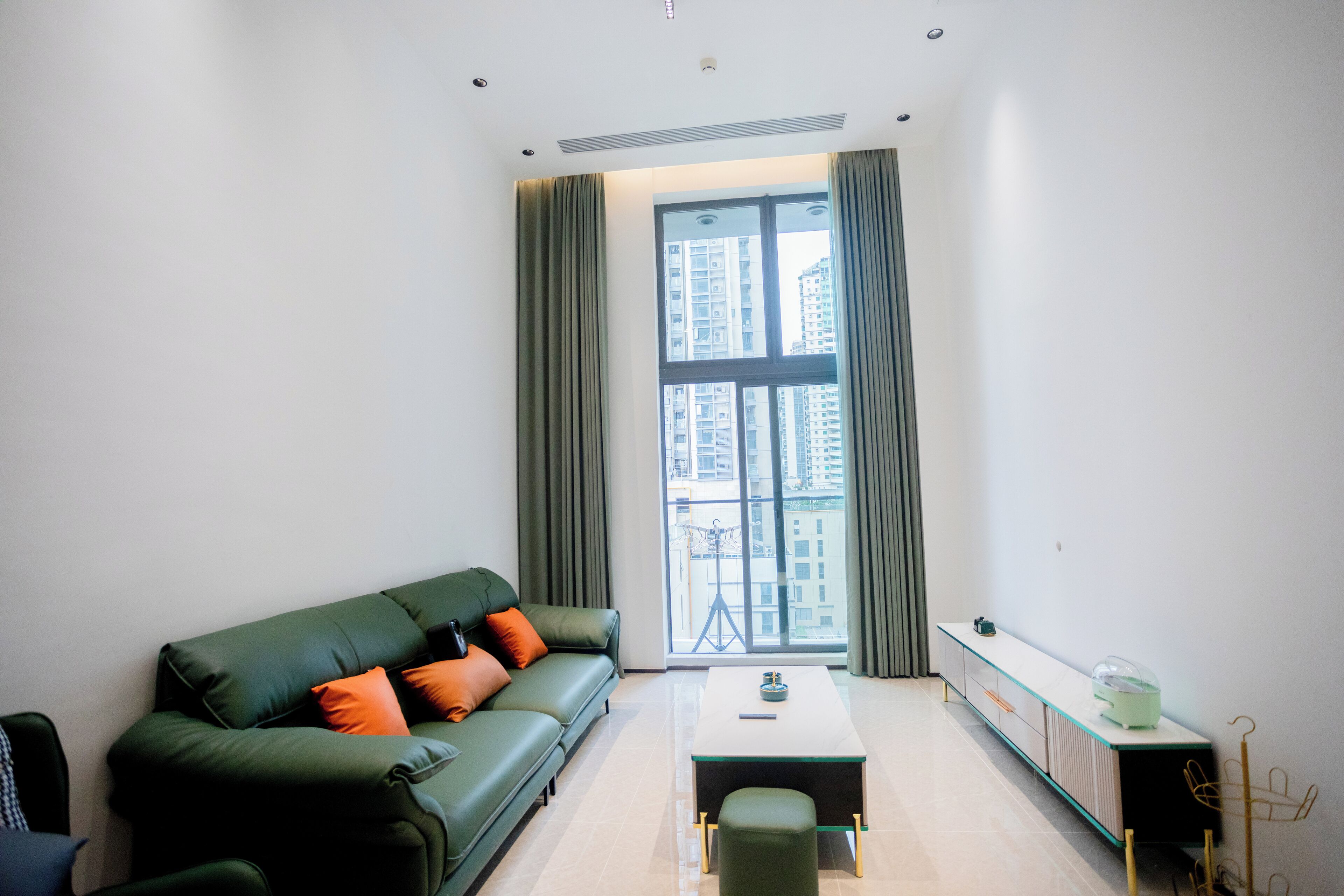 A modern living room with a green sofa, a small table, and a view of tall buildings through large windows.