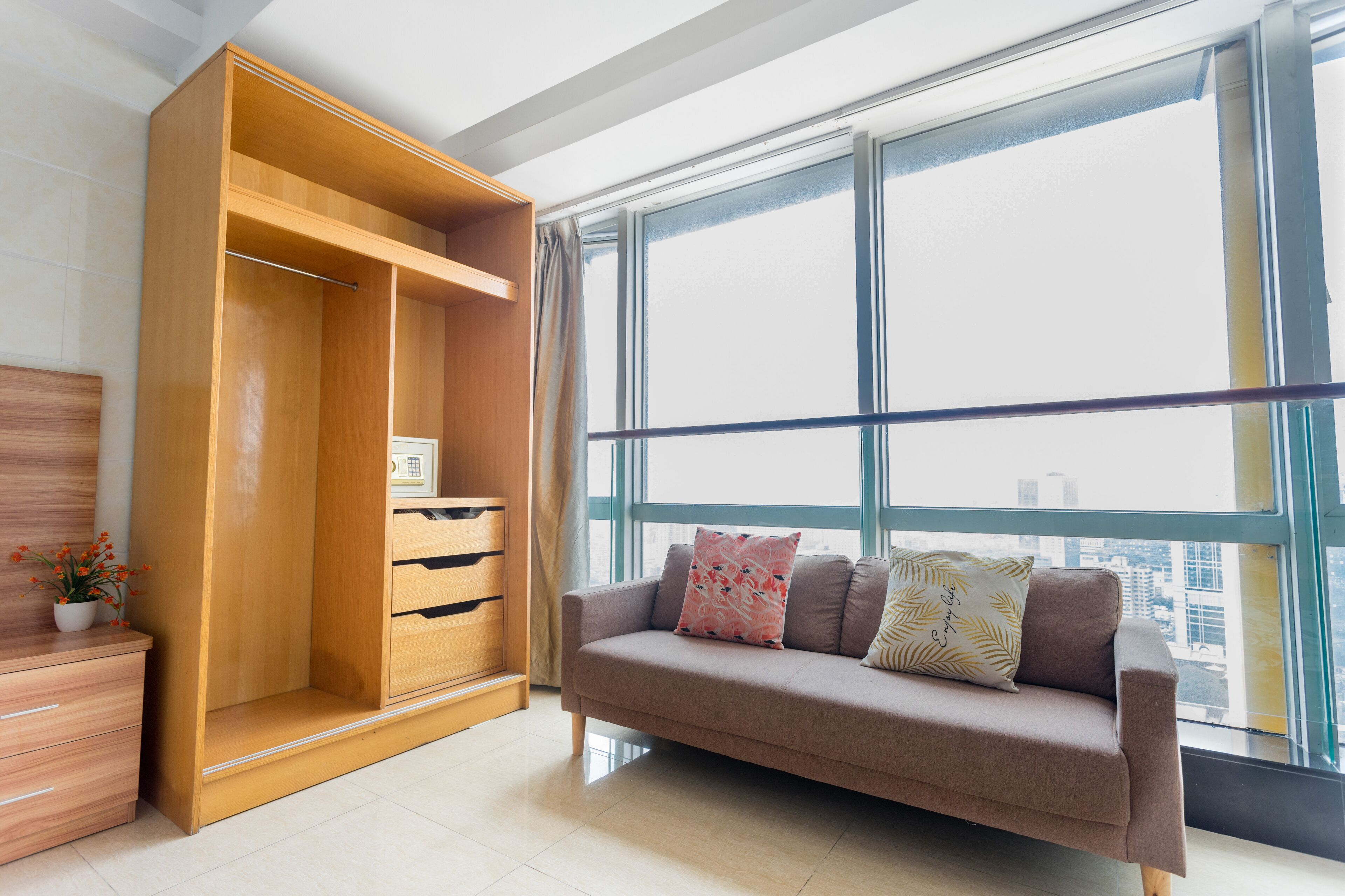 A modern living room with a large window, a wooden wardrobe, a sofa with cushions, and a small table with a plant.