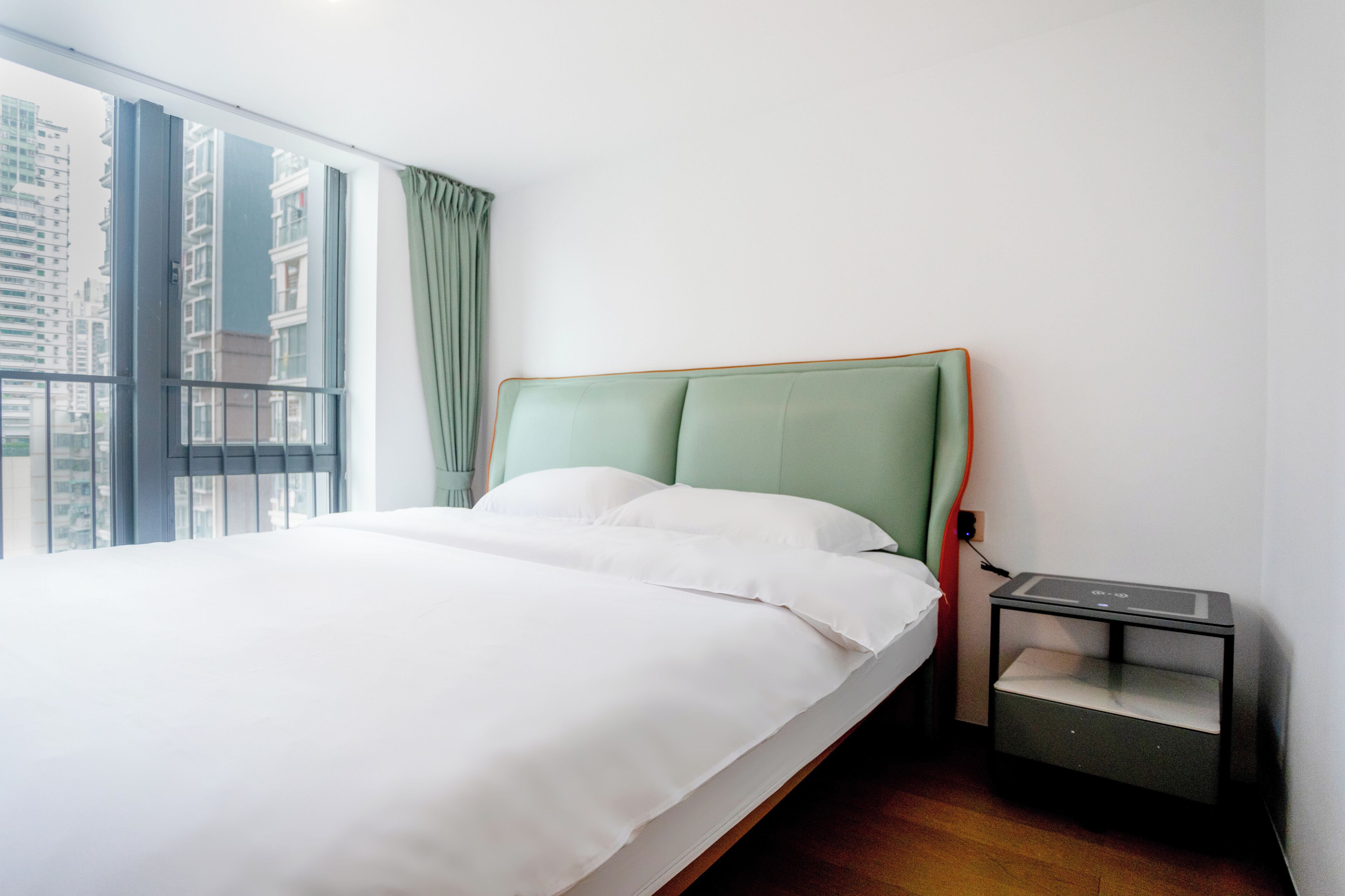 A neatly made bed with white linens, a green headboard, and a bedside table with a charging cable.