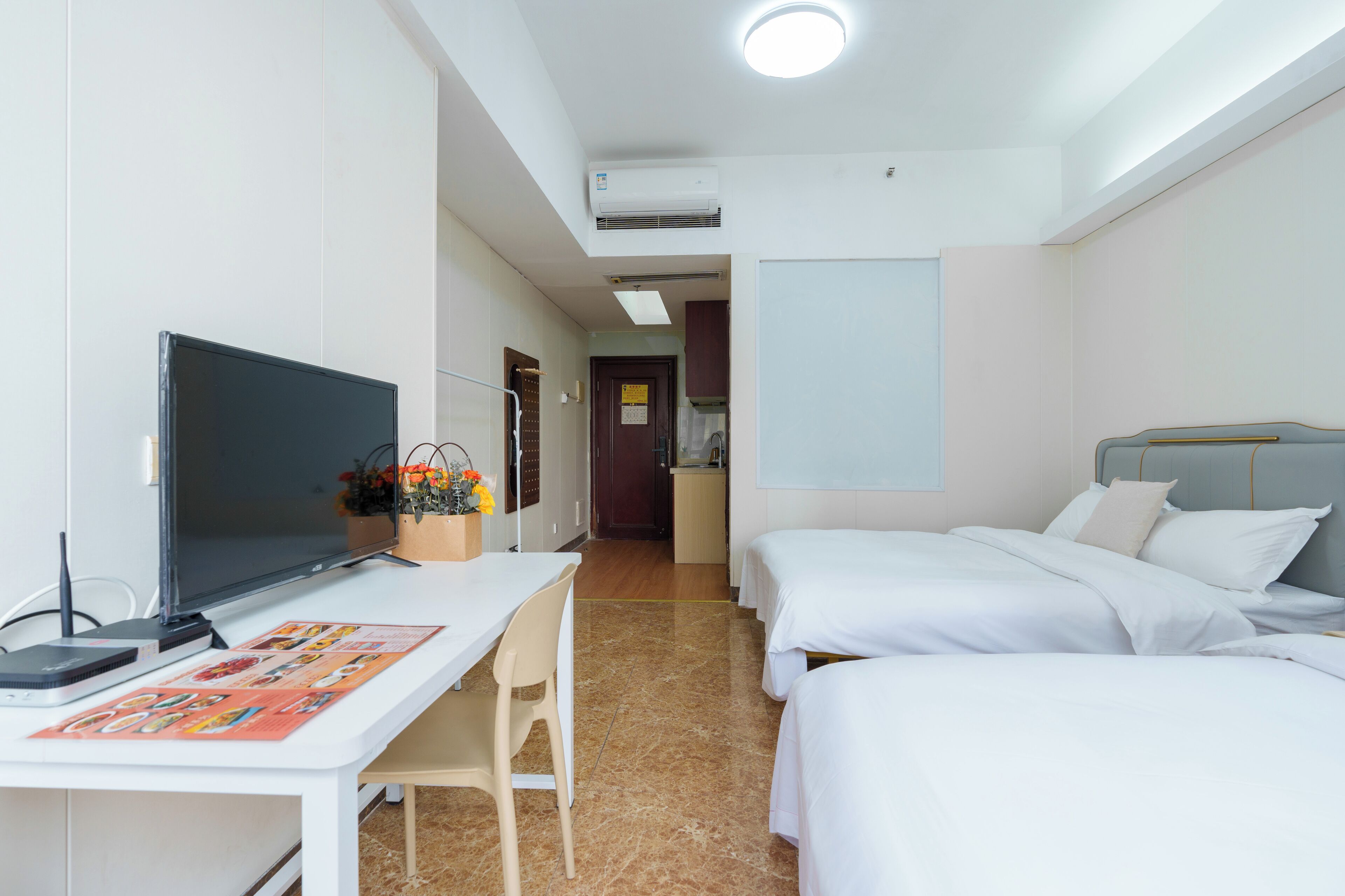 A hotel room with a desk, two beds, a TV, and a small kitchenette.