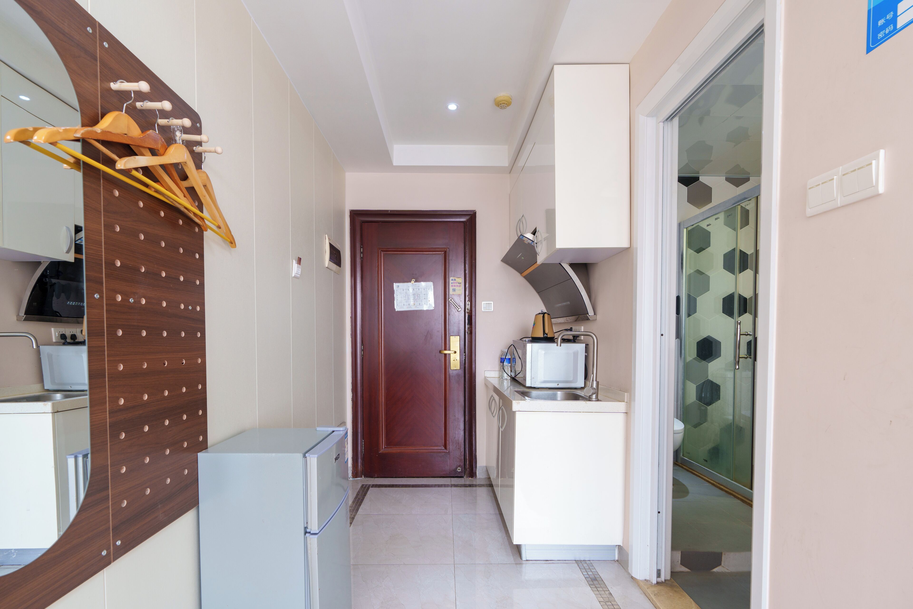 Un couloir étroit avec un étendoir en bois fixé au mur, un petit réfrigérateur, un four à micro-ondes posé sur un meuble blanc et une douche en verre dont le sol est recouvert de carreaux hexagonaux.