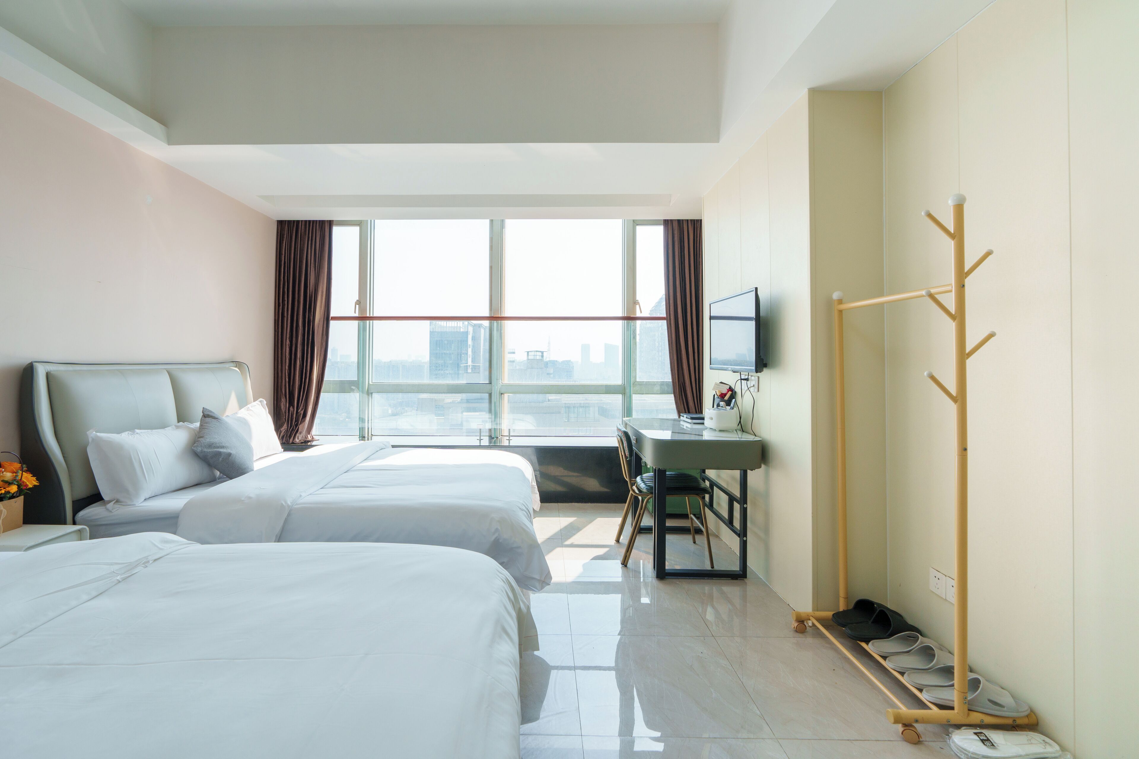 Quarto de hotel com duas camas, uma escrivaninha, um cabideiro e uma janela grande com vista para a cidade.