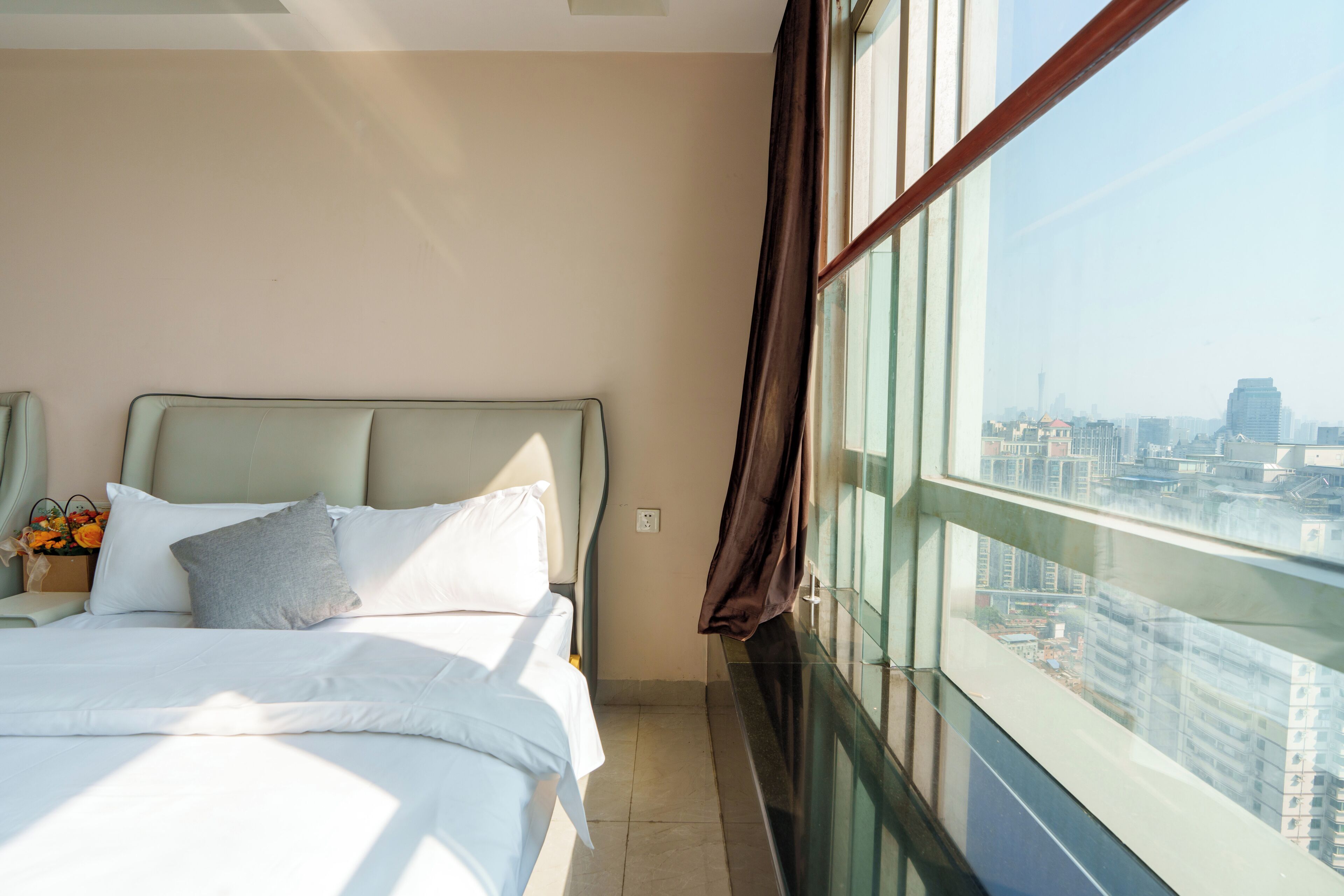 A hotel room with a bed, a nightstand with a lamp, and a view of the city through large windows.
