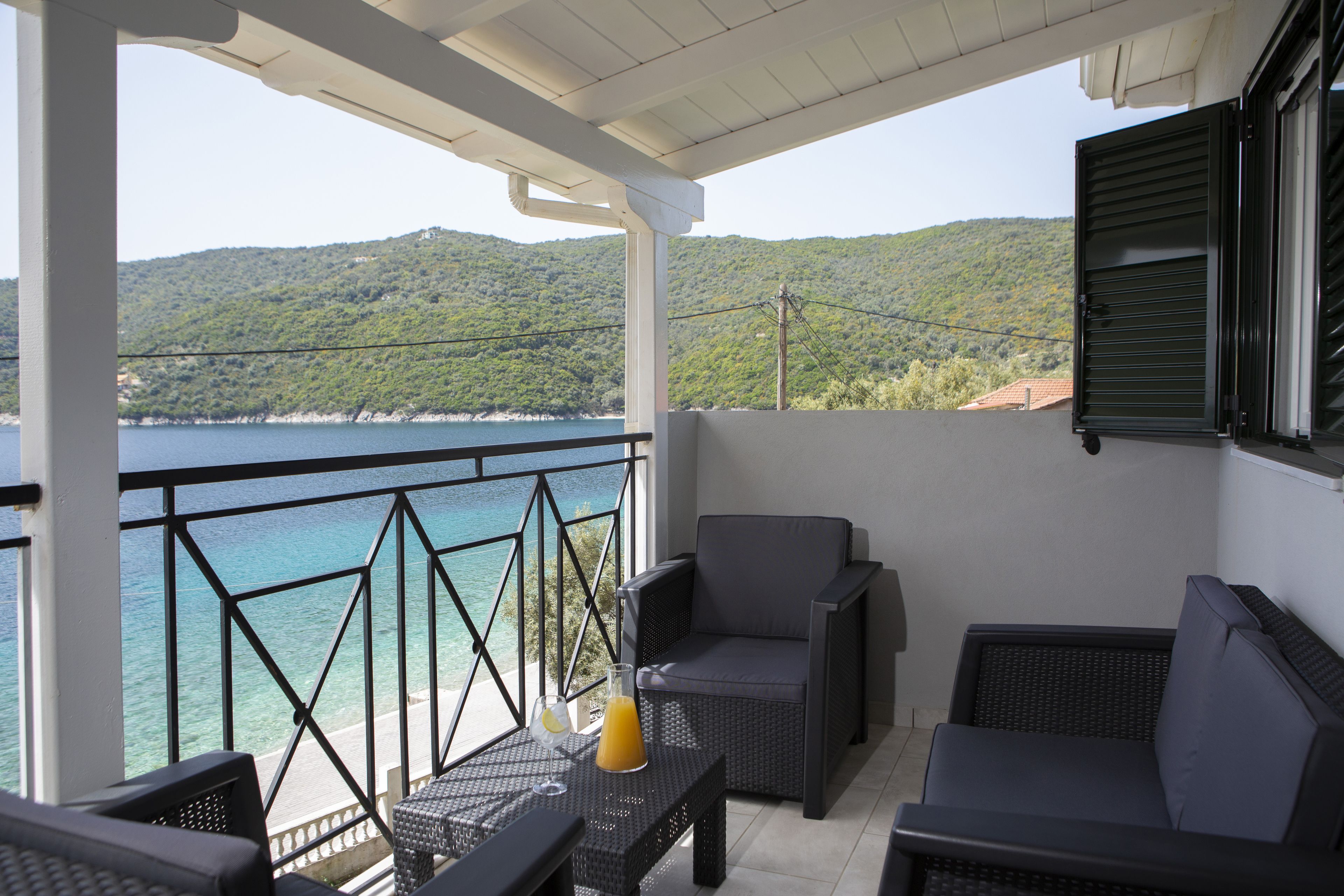 A balcony with wicker furniture, a table with a drink, and a view of the sea and hills.