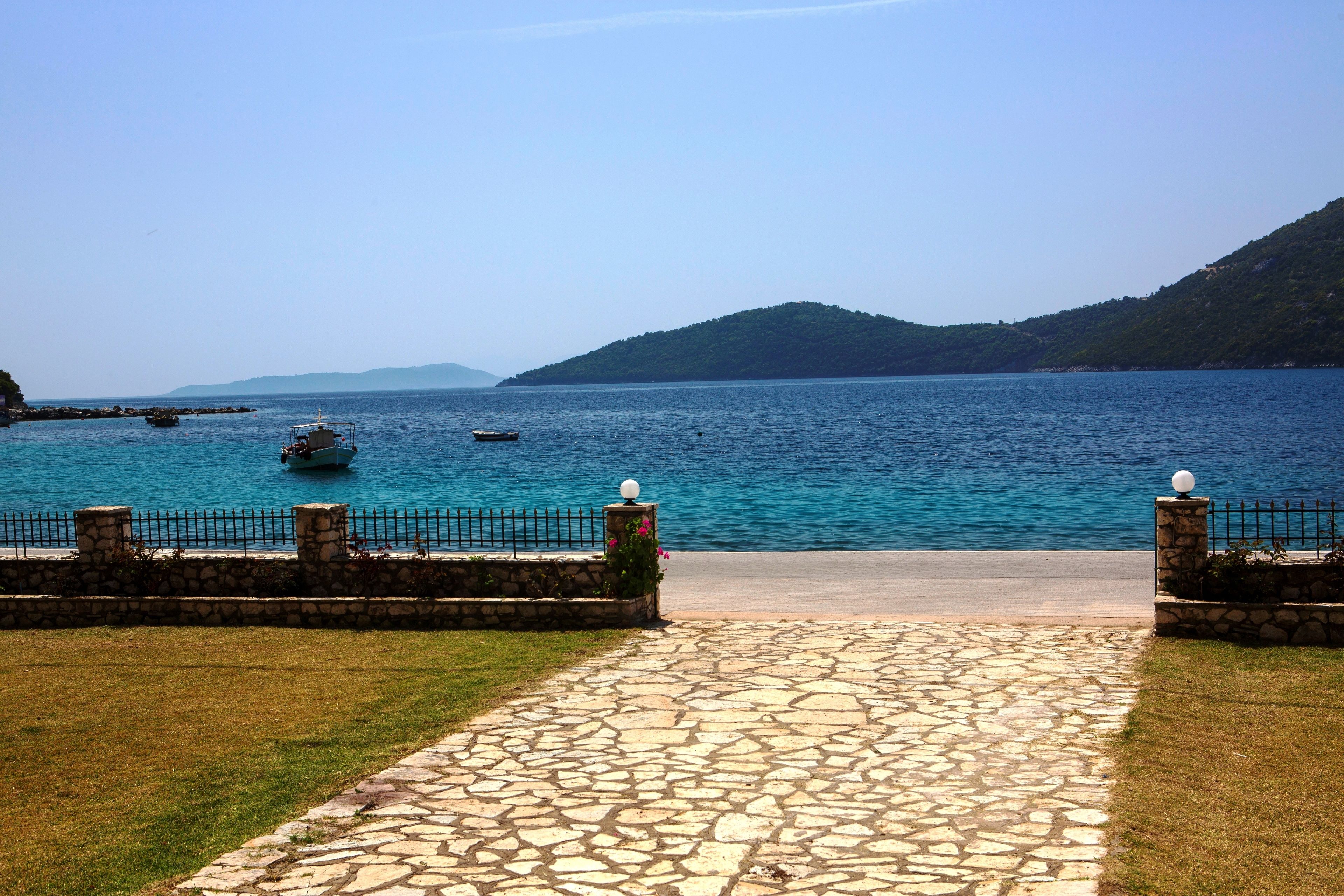 A stone pathway leading to a coastal view with a clear blue sea, boats, and a mountainous backdrop.