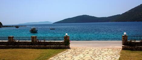 A stone pathway leading to a coastal view with a clear blue sea, boats, and a mountainous backdrop.