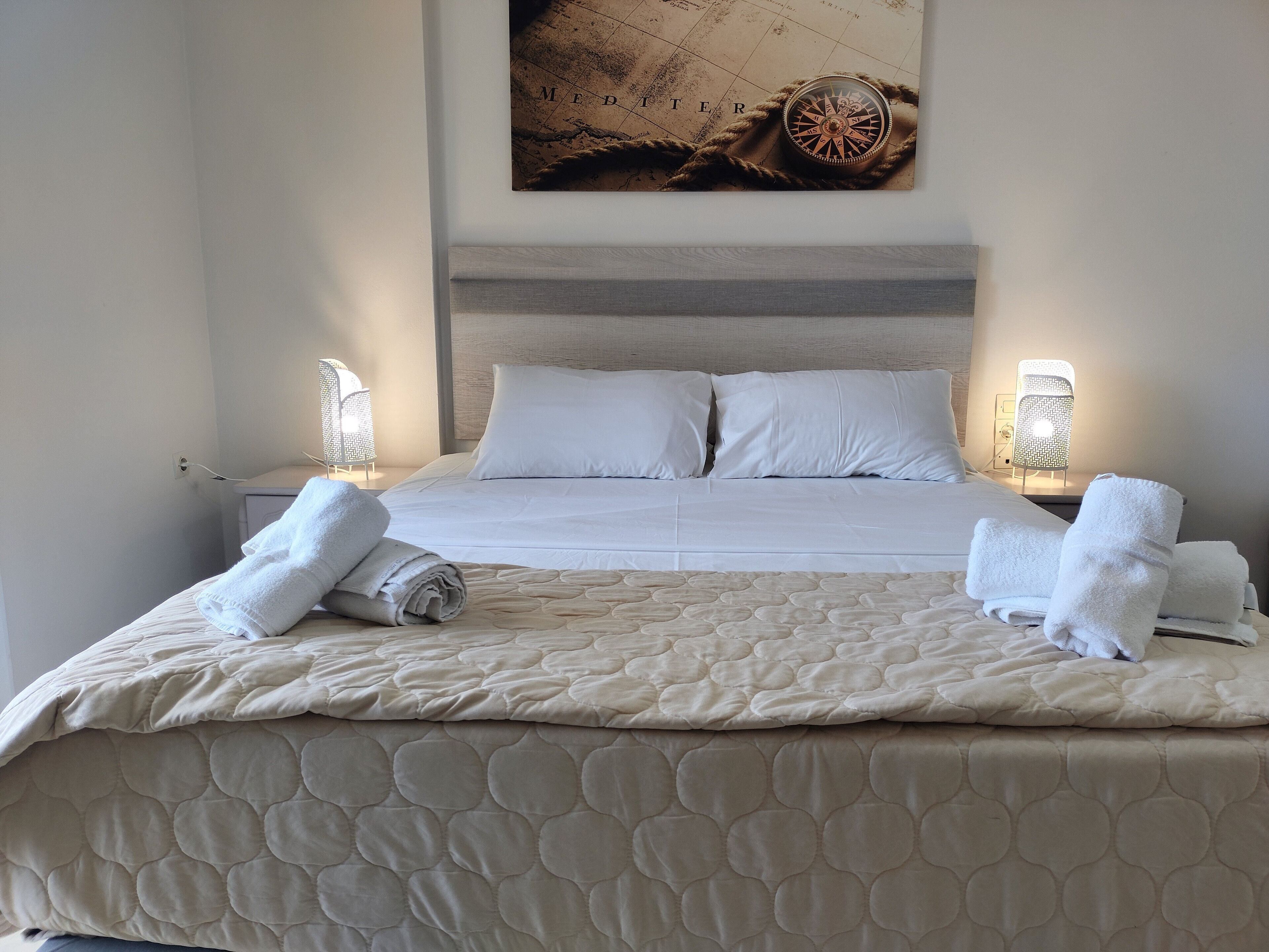 A neatly made bed with white linens and a quilted duvet, two bedside lamps, and a wall-mounted compass artwork.