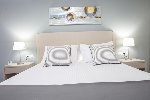 Een net opgemaakt bed met gestreepte kussens, twee nachtkastjes met lampen, en een abstract schilderij aan de muur.