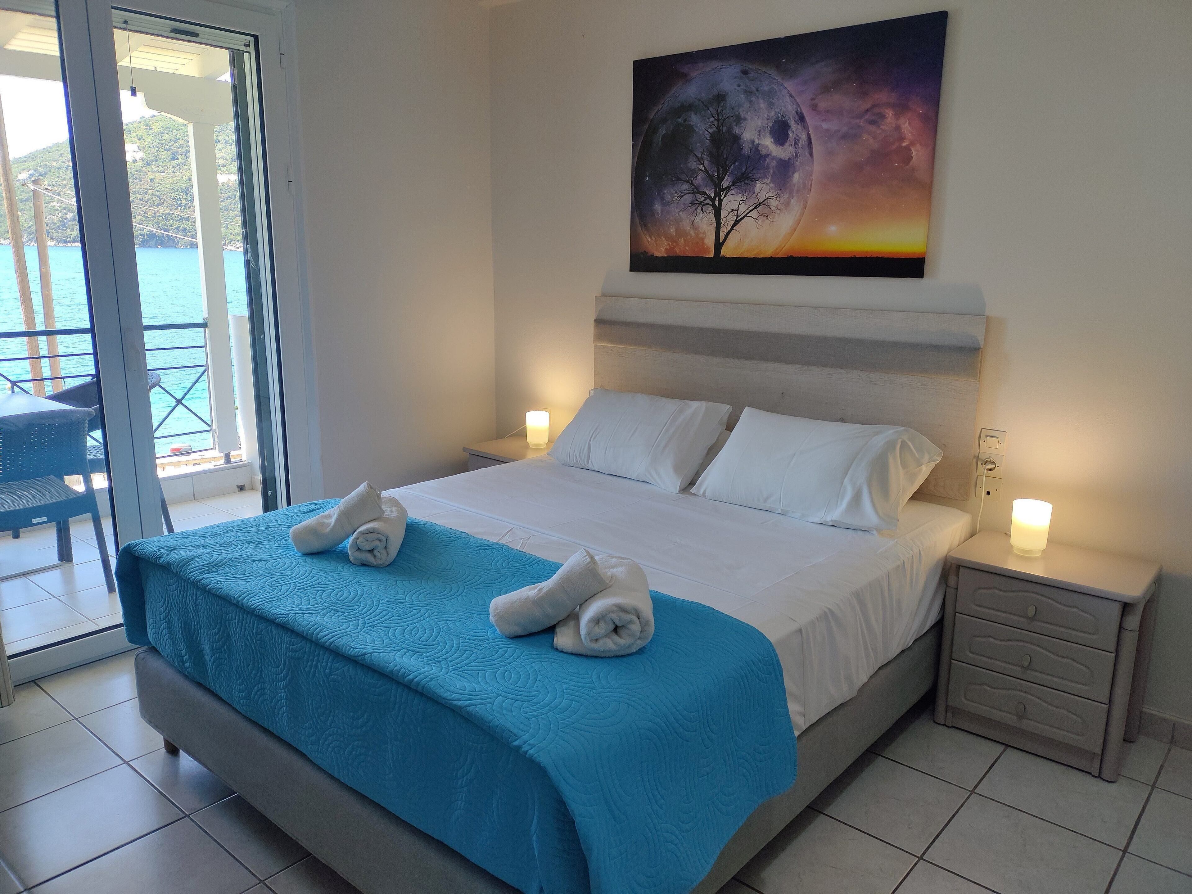 A hotel room with a bed, a nightstand, a painting of a tree and moon, and a view of the sea through a glass door.