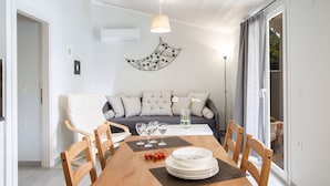 A dining area with a wooden table set for four, surrounded by chairs. A sofa with striped cushions is in the background, and a wall-mounted crescent moon decoration is visible. - Captain Nick (Lefkada)