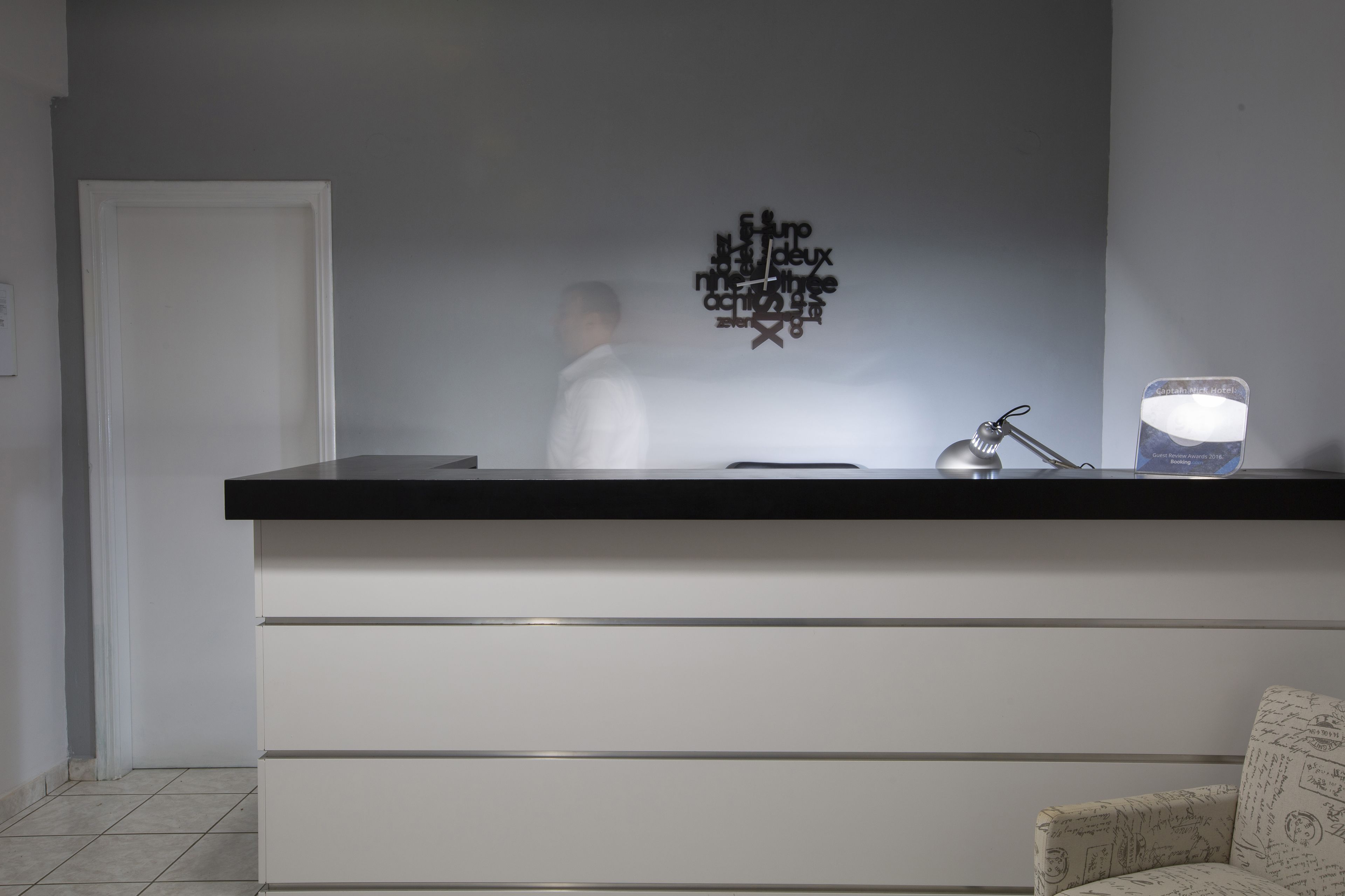 A reception desk with a black top and white drawers, a wall-mounted sign with text, a lamp, and a chair with a patterned cushion.