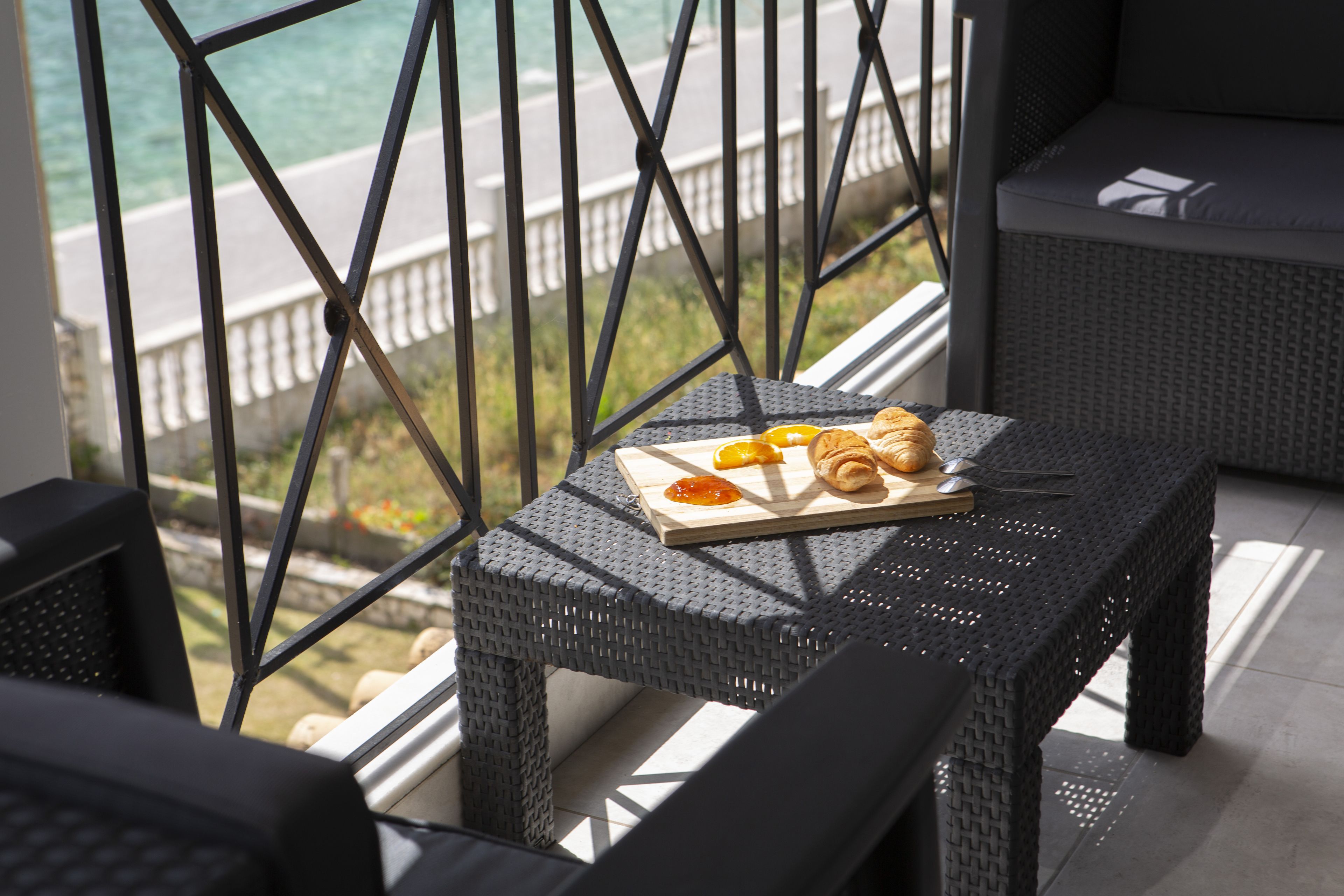 Una mesa de mimbre con un plato de cruasanes y rodajas de naranja, una computadora portátil y un teléfono inteligente en un balcón con vistas a un cuerpo de agua.