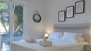 A neatly made bed with white linens, a bedside table with a lamp, and a view of a balcony with palm trees. - Captain Nick (Lefkada)