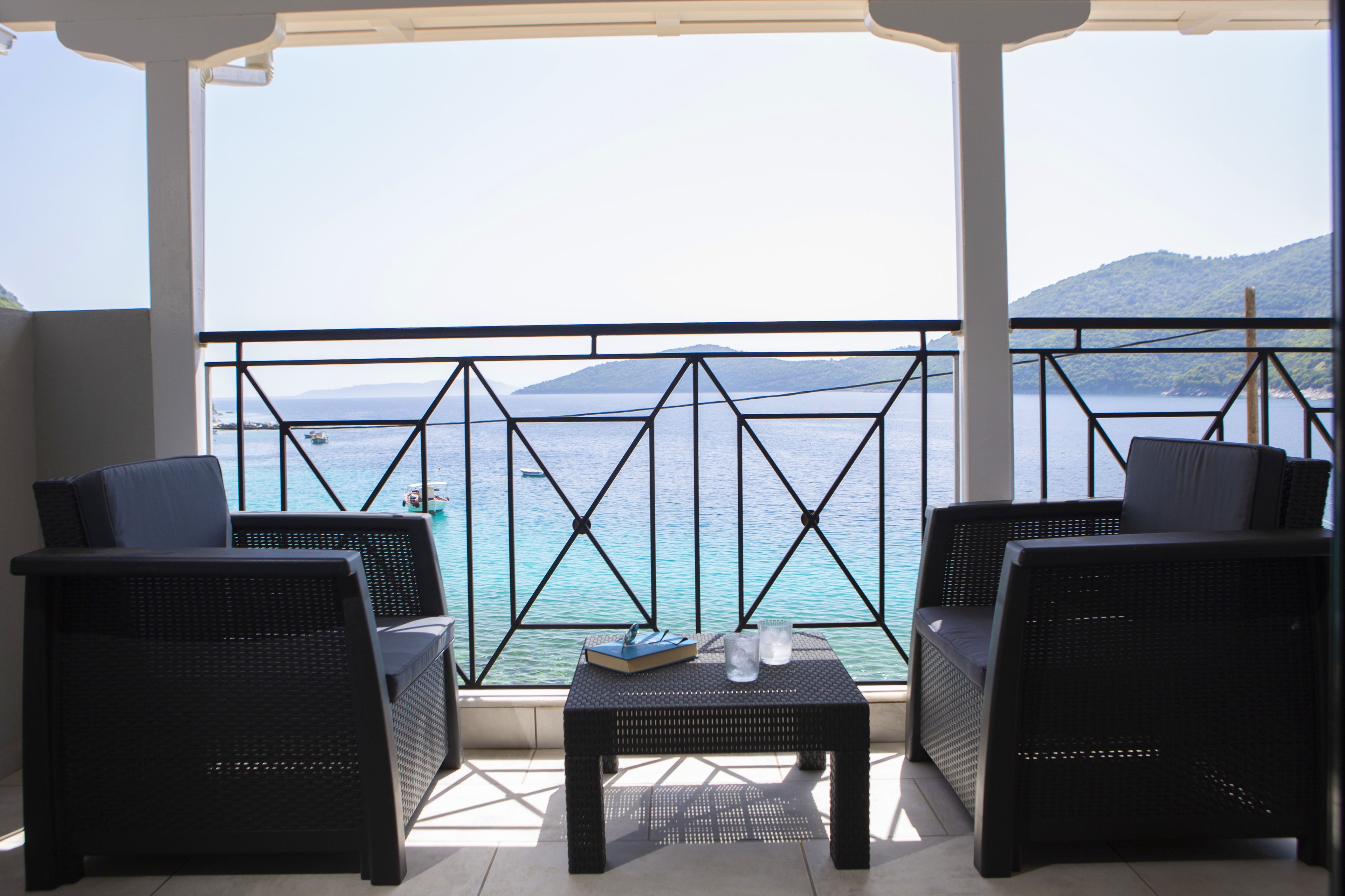 A balcony with wicker furniture and a table, overlooking a body of water and distant hills.