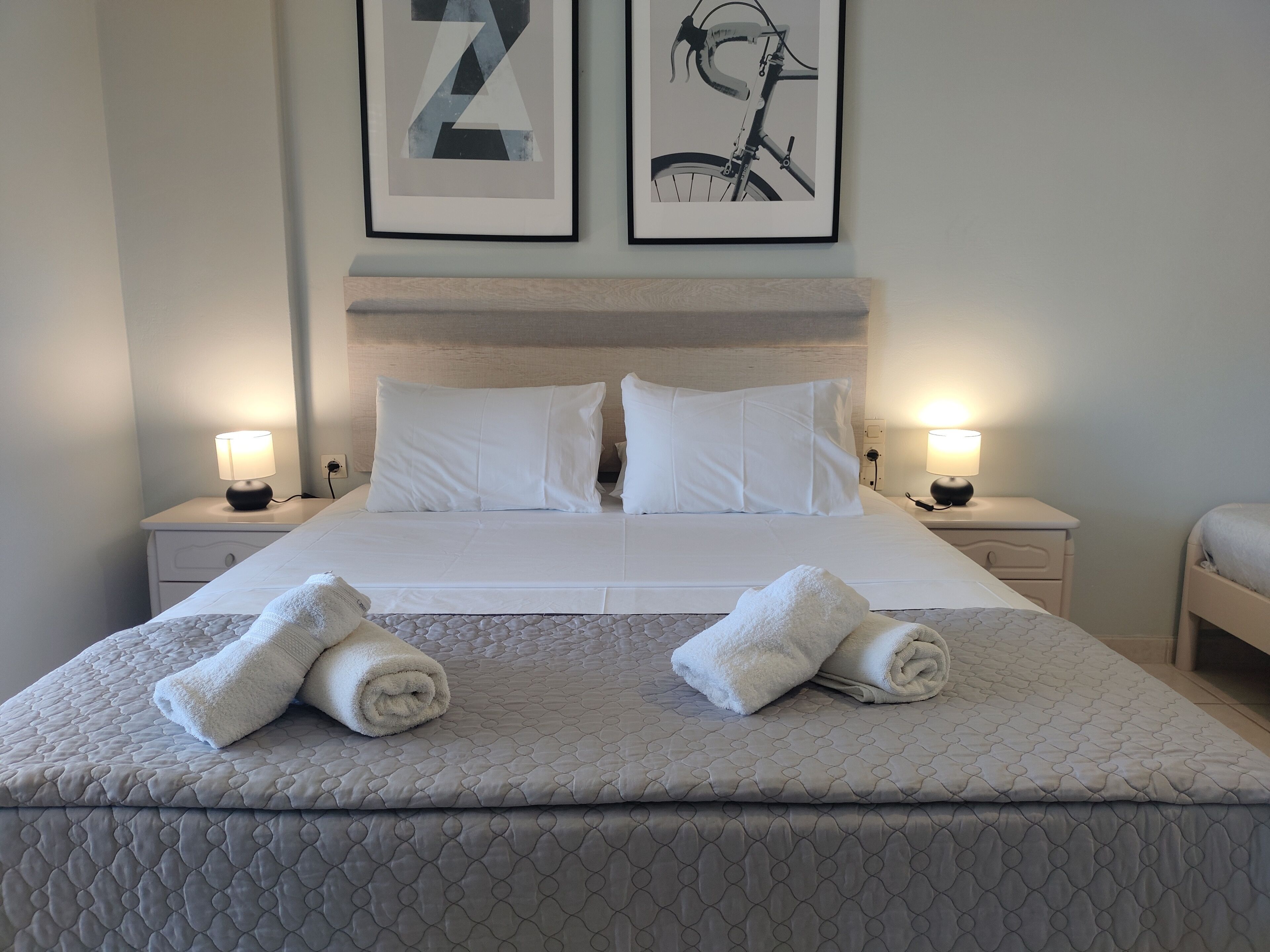 Uma cama bem arrumada com duas toalhas enroladas, roupa de cama branca e duas obras de arte emolduradas na parede.