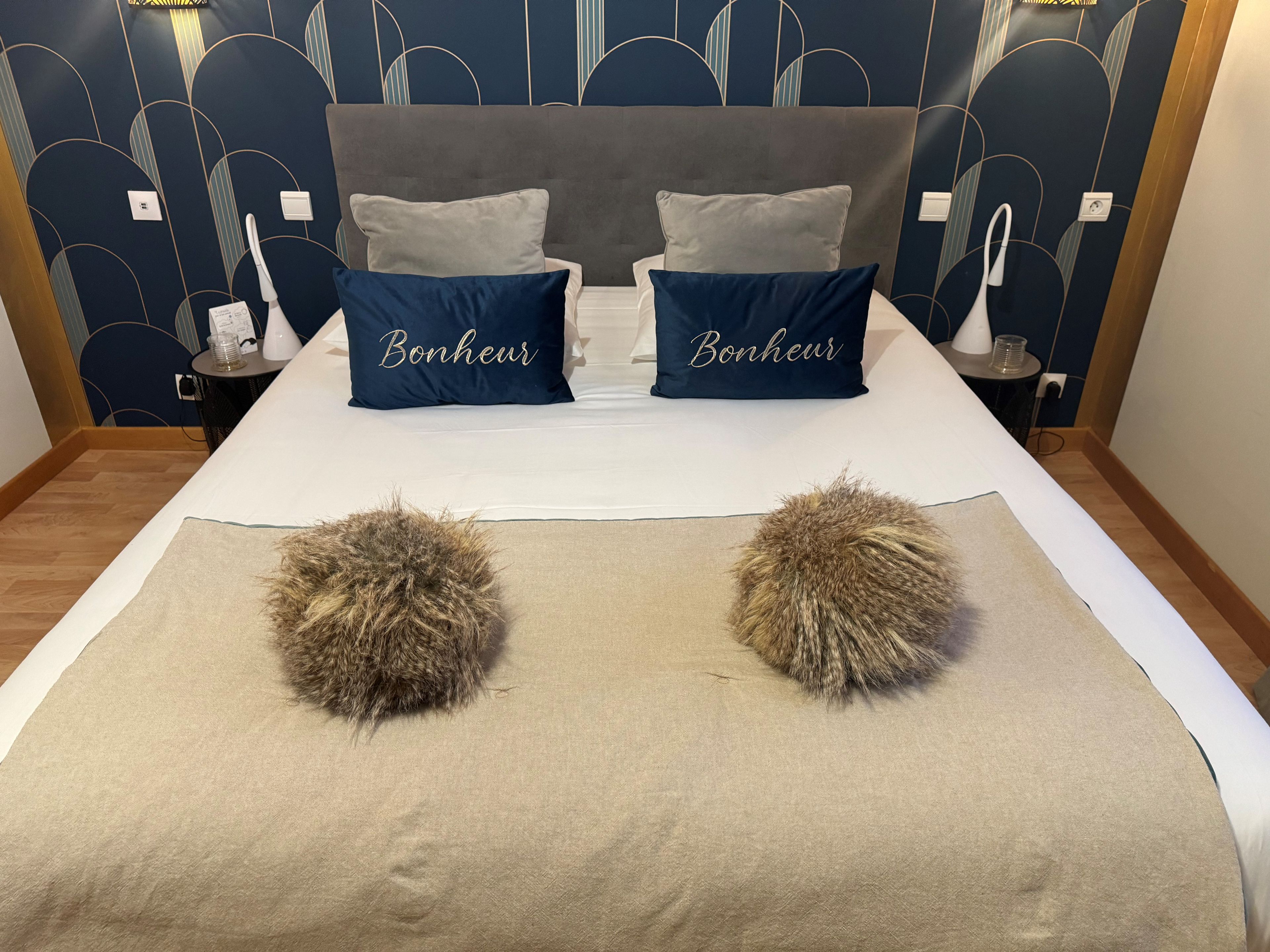 A bed with two pillows labeled 'Bonjour' and a beige throw blanket with two fluffy pom-poms.