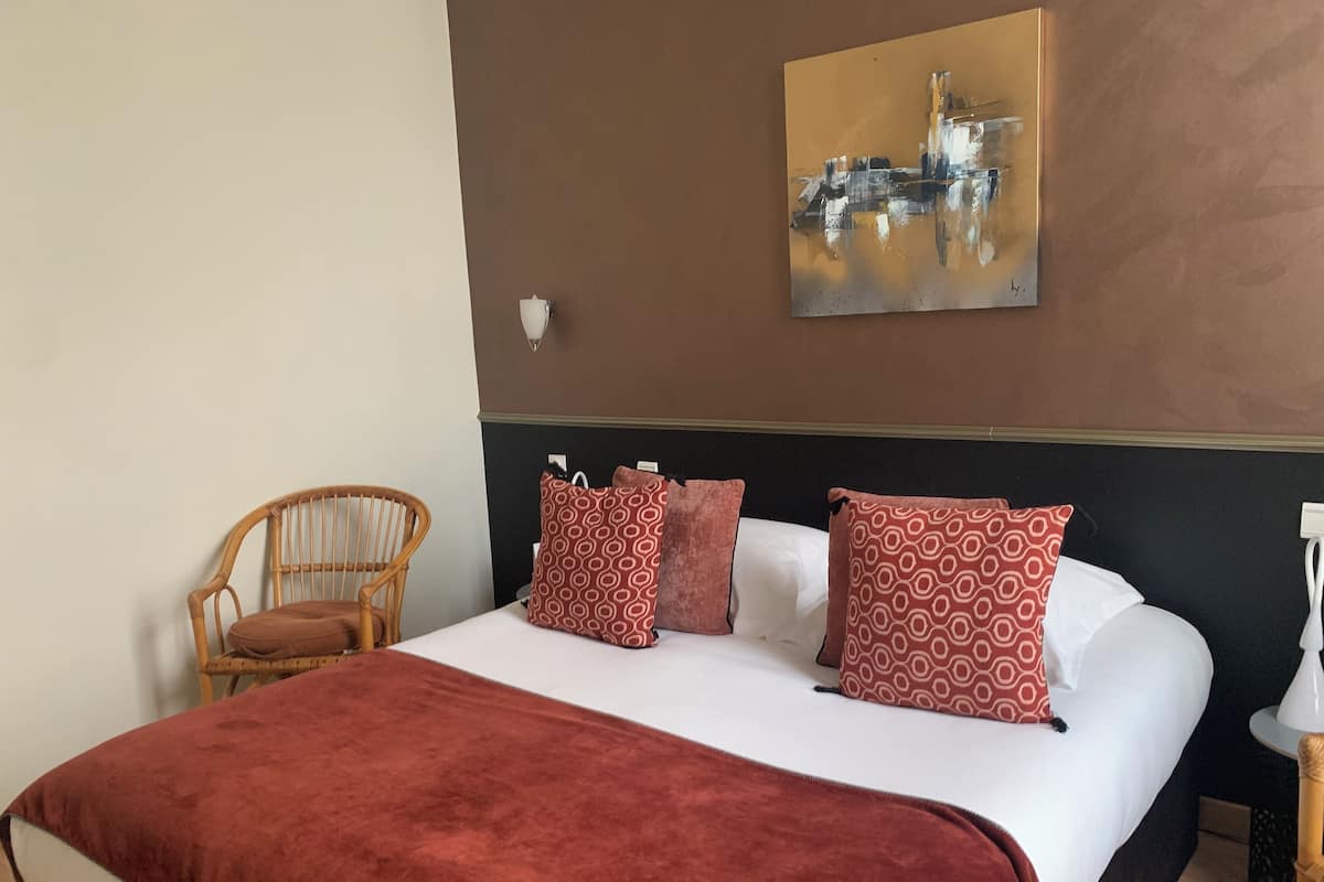 A neatly made bed with a red blanket, a wooden chair, patterned pillows, and a wall-mounted painting.