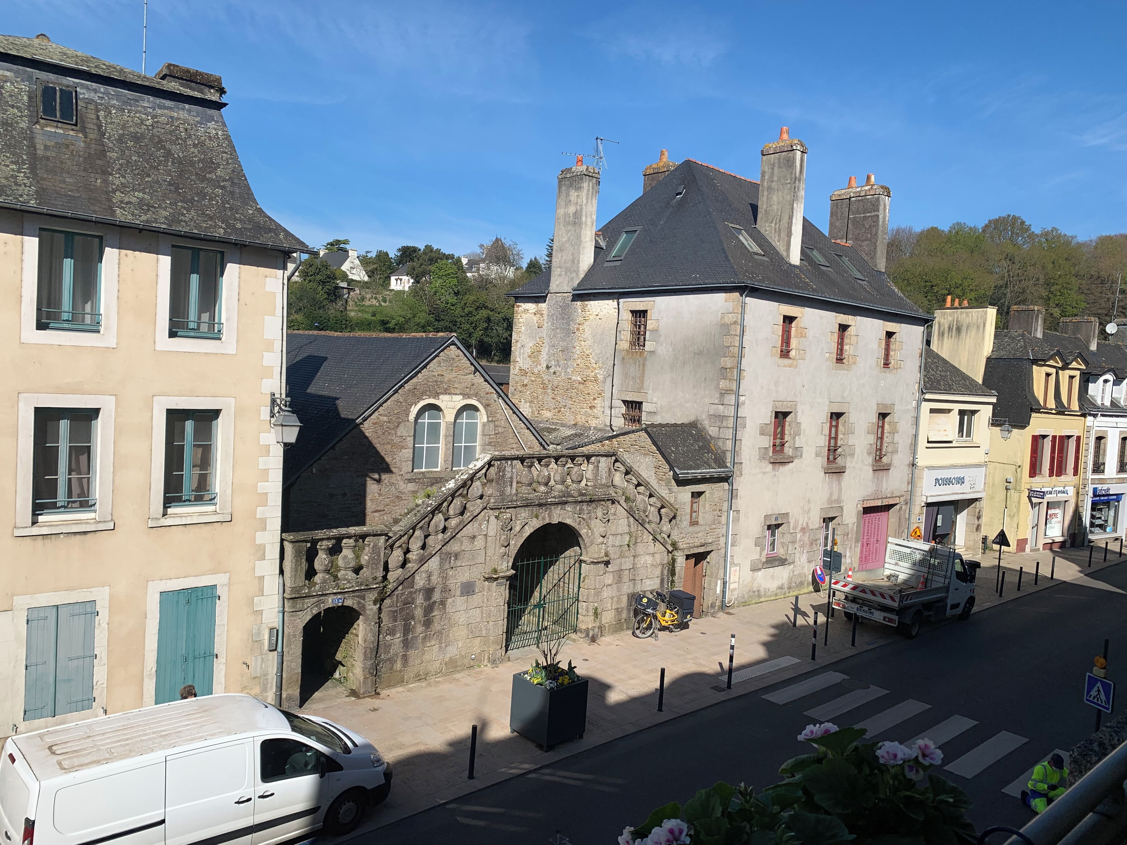 Une vue de rue avec un fourgon blanc garé sur le bord, un pont de pierre et des bâtiments dotés de différentes devantures de magasins.