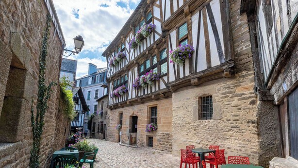 A narrow cobblestone street with traditional half-timbered buildings, a stone wall on one side, and a small outdoor seating area with green and red chairs. - Hôtel Vintage (Quimperle)