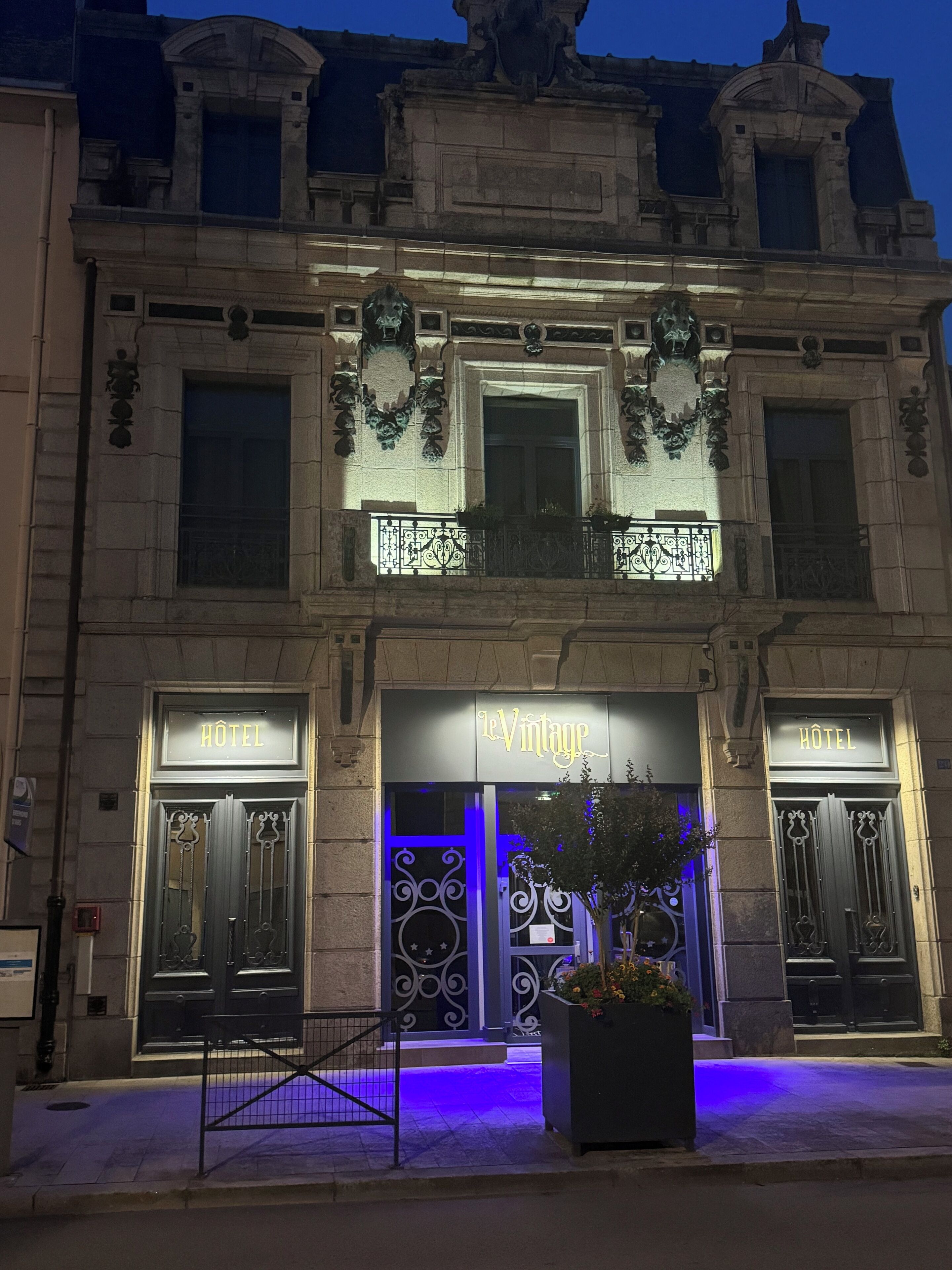 Fachada de hotel con intrincados detalles de hierro forjado, una entrada central con un cartel que dice "LeVintage" y una planta en maceta al frente.