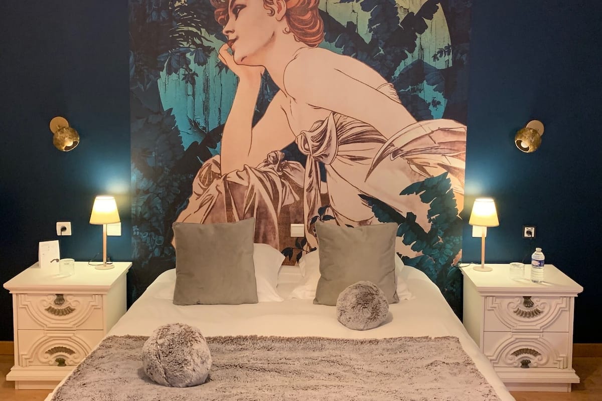 A bedroom with a large tapestry of a woman on the wall, two bedside tables with lamps, a bed with pillows, and a rug.