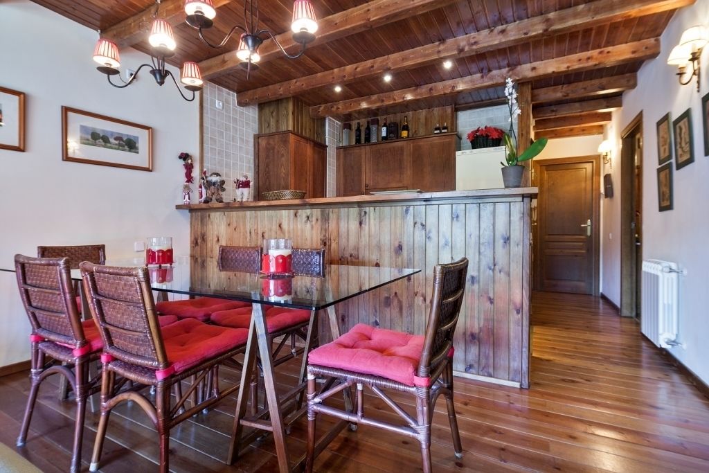A wooden interior with a glass dining table and red cushioned chairs, a wooden bar area, and framed pictures on the walls.