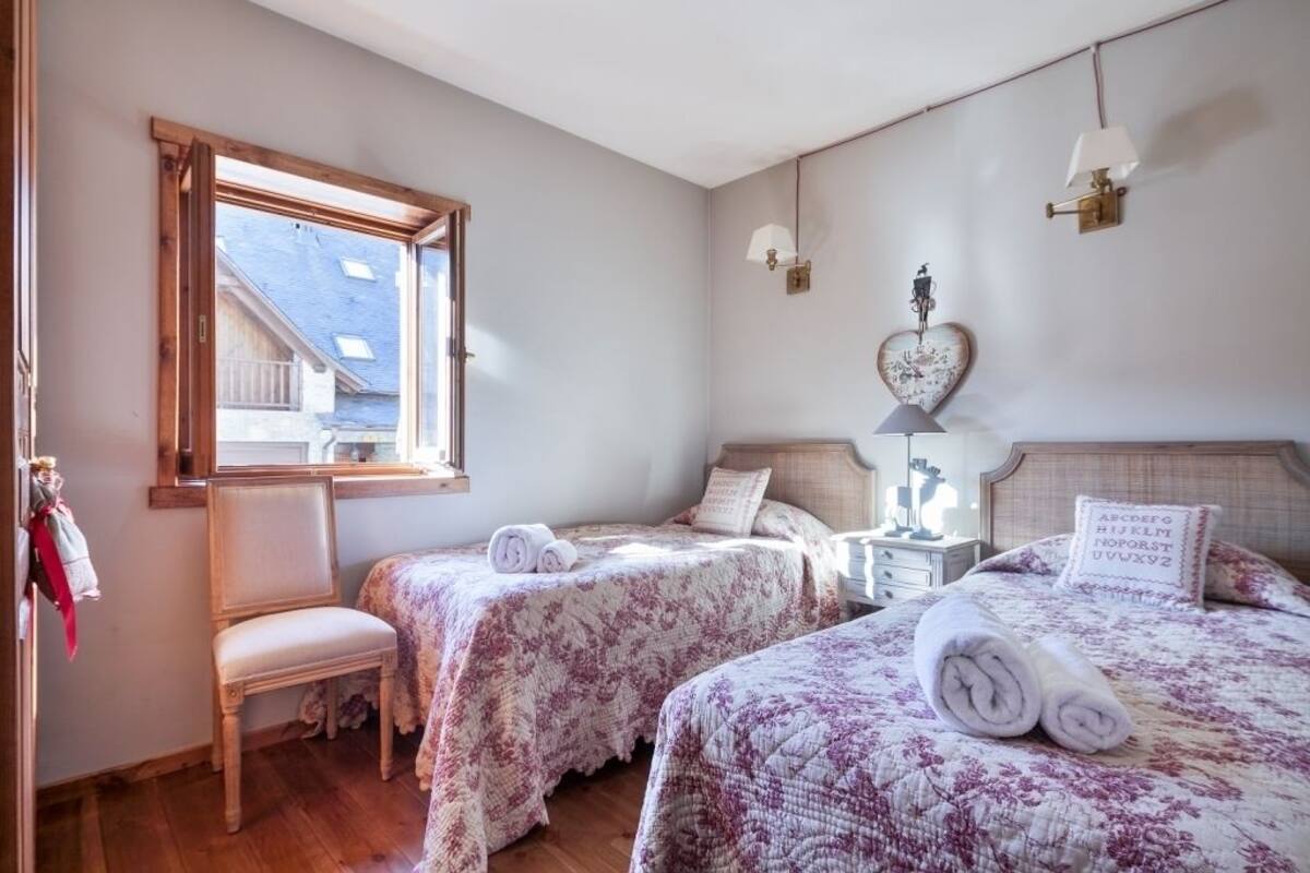 Ein Schlafzimmer mit zwei Betten, einem Holzstuhl, einem Nachttisch mit Lampe und einem Fenster mit Blick auf Gebäude.