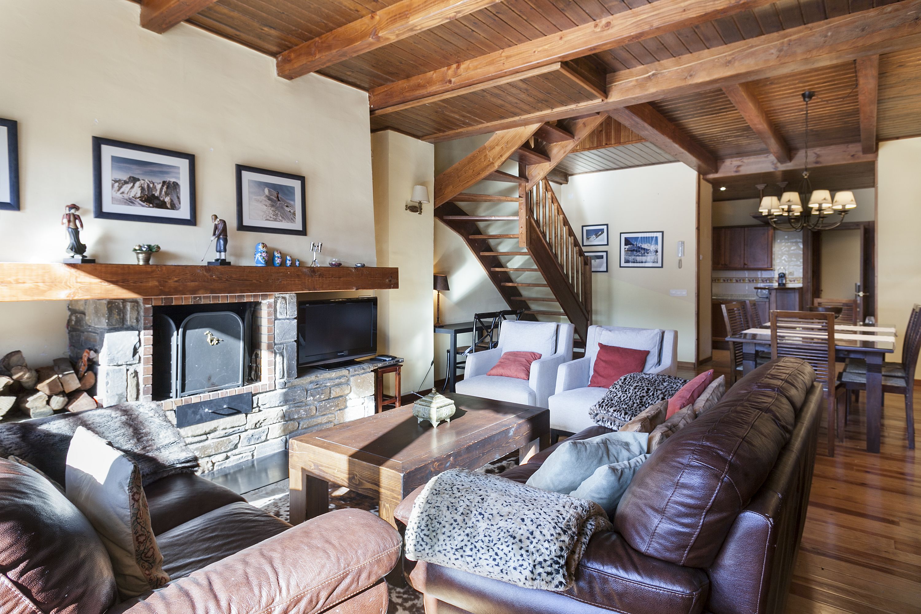 Ein gemütliches Wohnzimmer mit Kamin aus Stein, Ledersofas und einer Holztreppe, die in einen Essbereich führt.