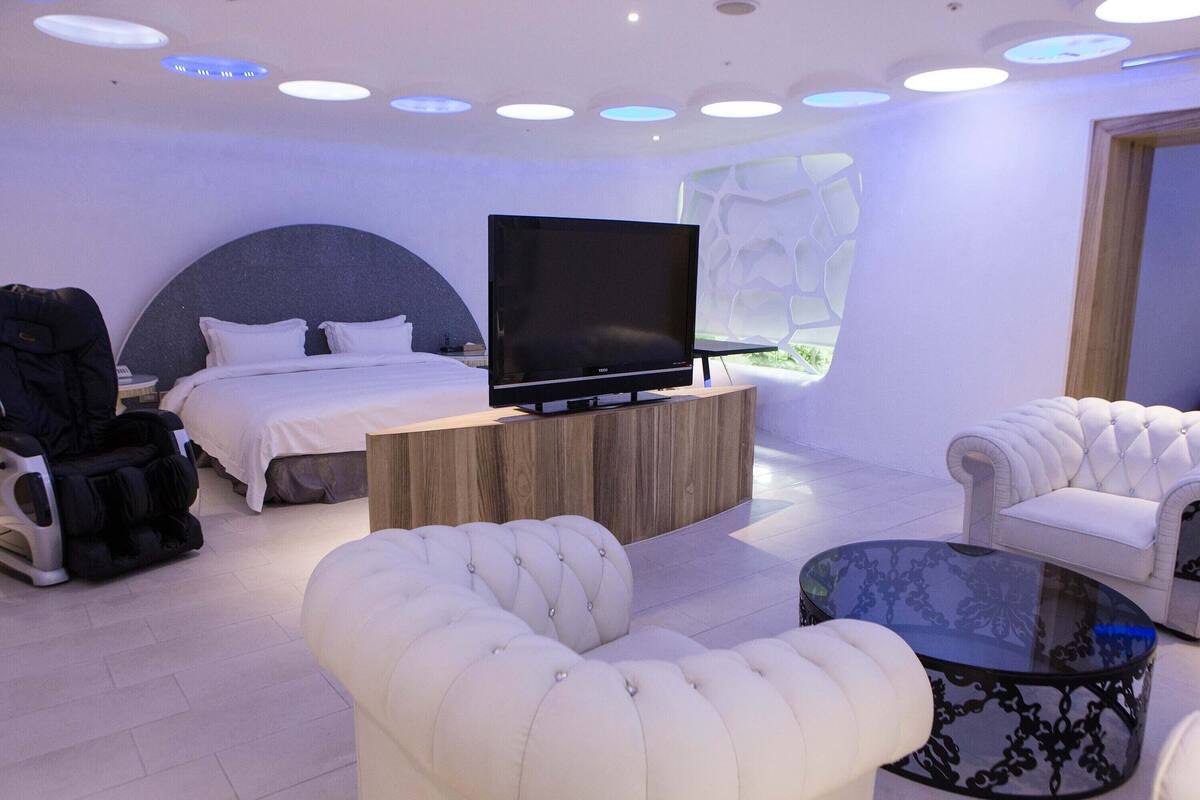 Ein modernes Hotelzimmer mit einem großen Bett, einem Flachbildfernseher, einem Massagesessel, einem weißen Sofa und einem Glastisch.