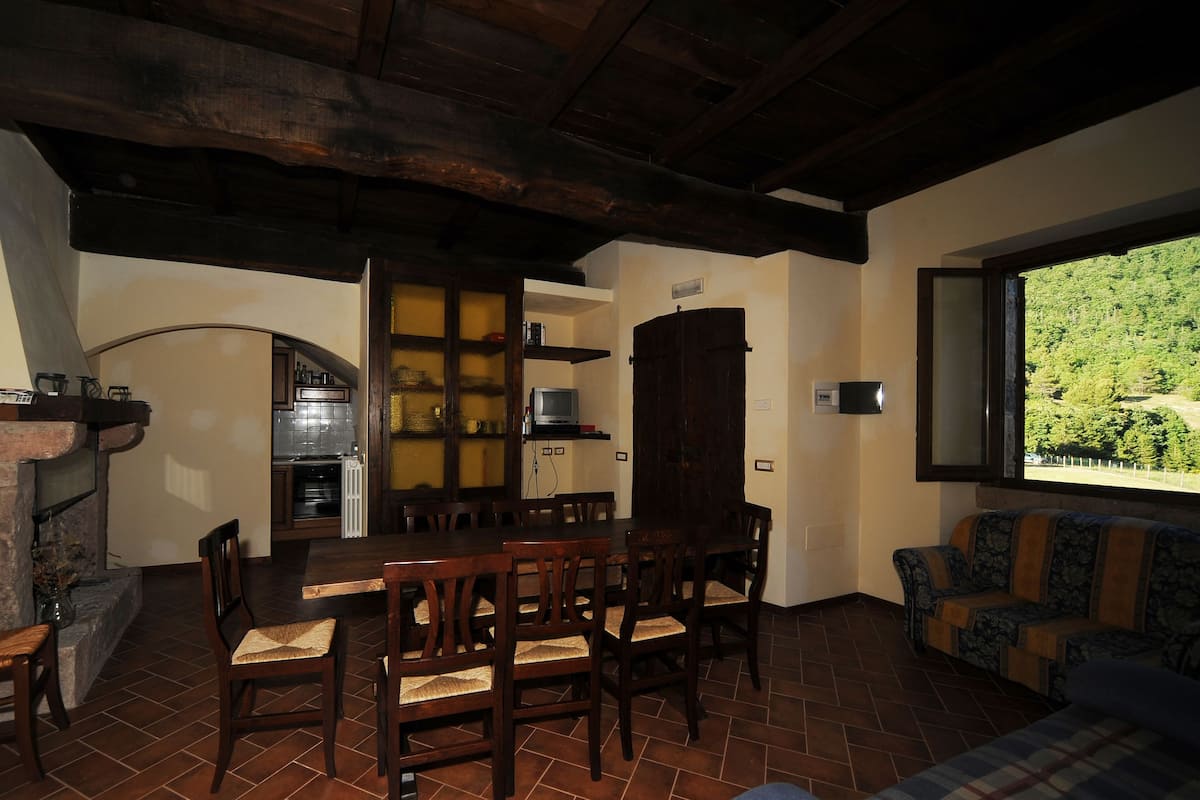 Uma área de jantar acolhedora com piso de tijolos, cadeiras de madeira, lareira e janela com vista para o verde.