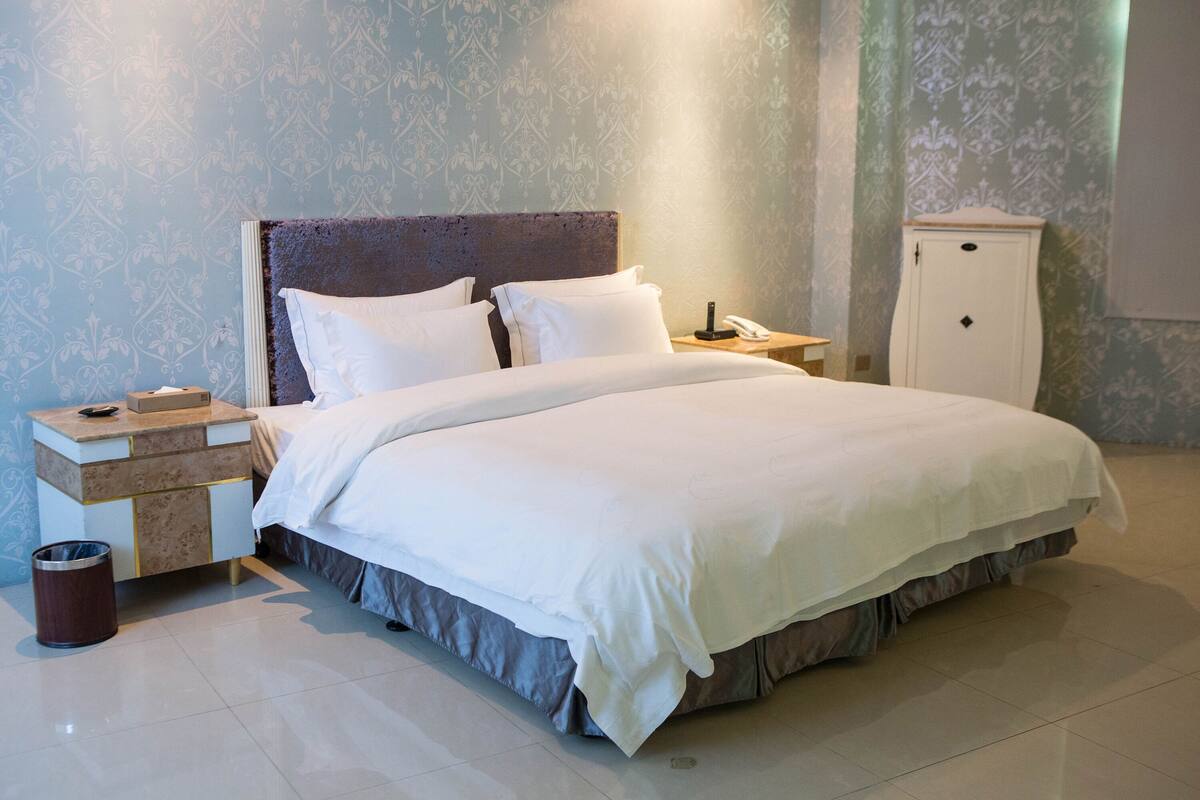 A neatly made bed with white linens, a bedside table with a lamp, and a small cabinet.