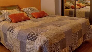 Una cama bien tendida con ropa de cama estampada y dos almohadas naranjas, un cabecero de madera y un espejo que refleja la habitación.