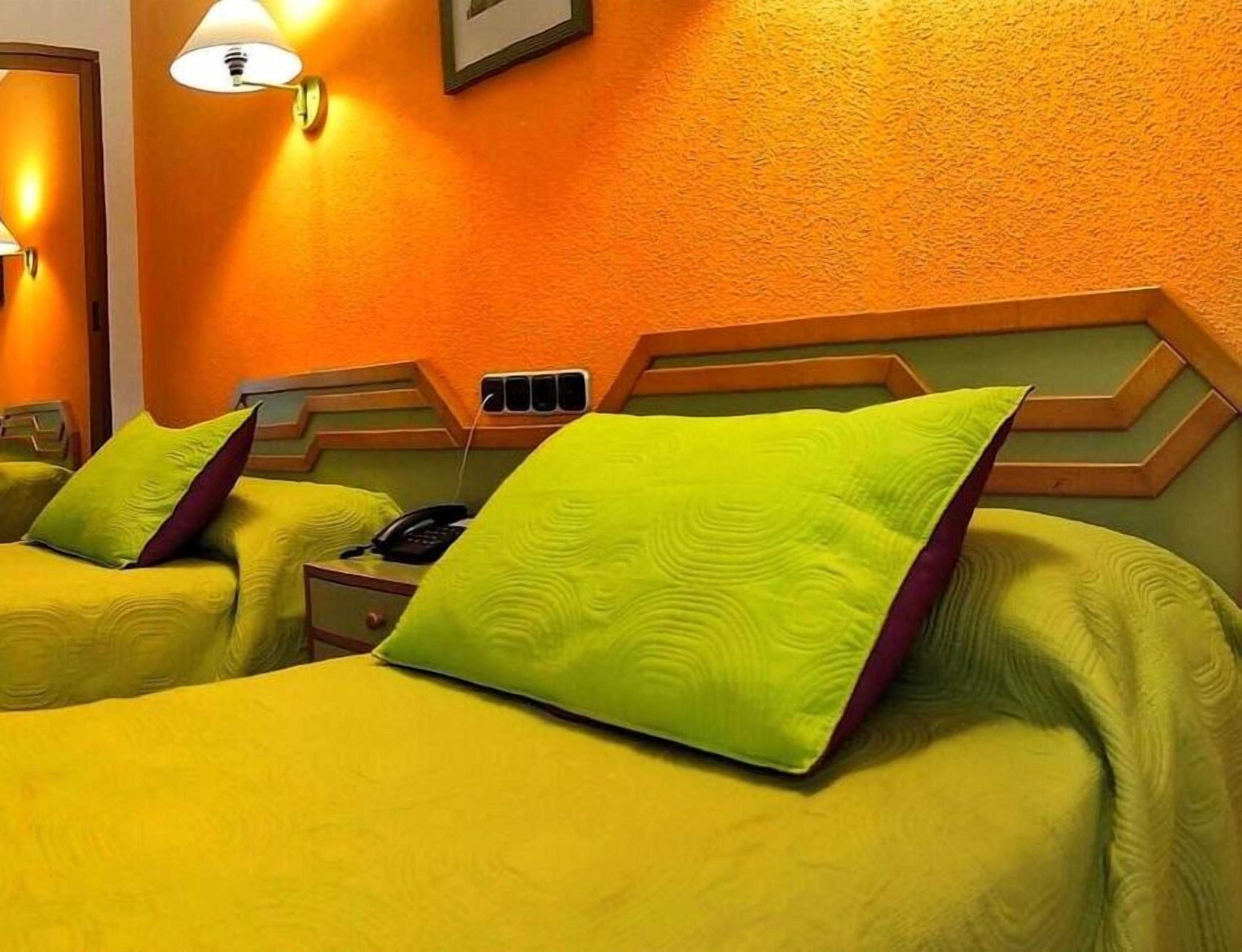 A hotel room with two beds, each with a green pillow and a headboard with a zigzag pattern. There is a bedside table with a phone and a lamp.
