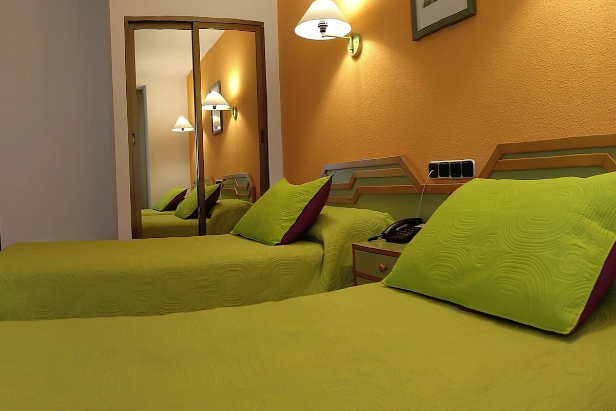 A hotel room with two beds, each with green bedding and pillows, a bedside telephone, and wall-mounted lamps.