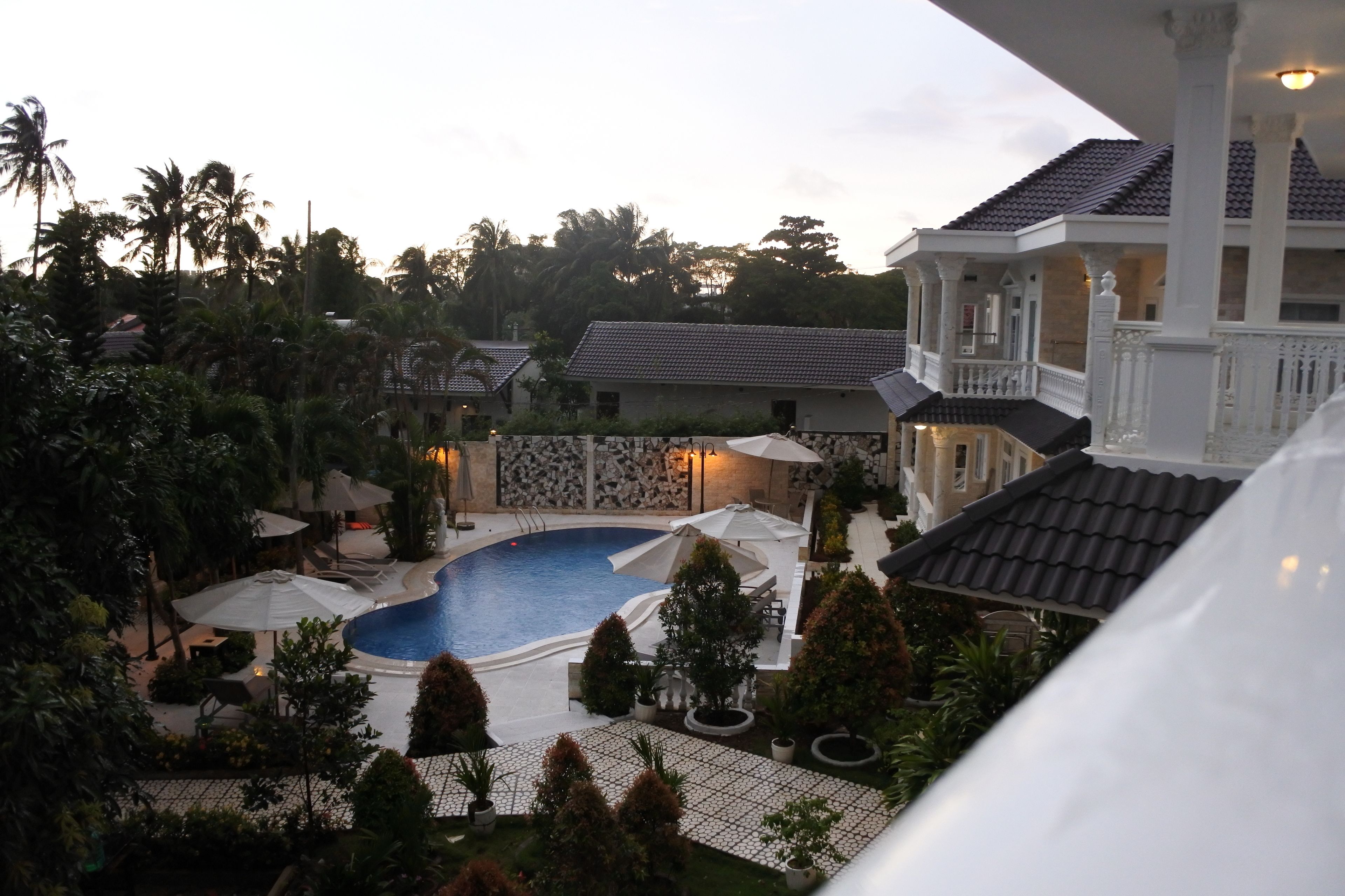 A swimming pool surrounded by umbrellas and potted plants, with a large building in the background.