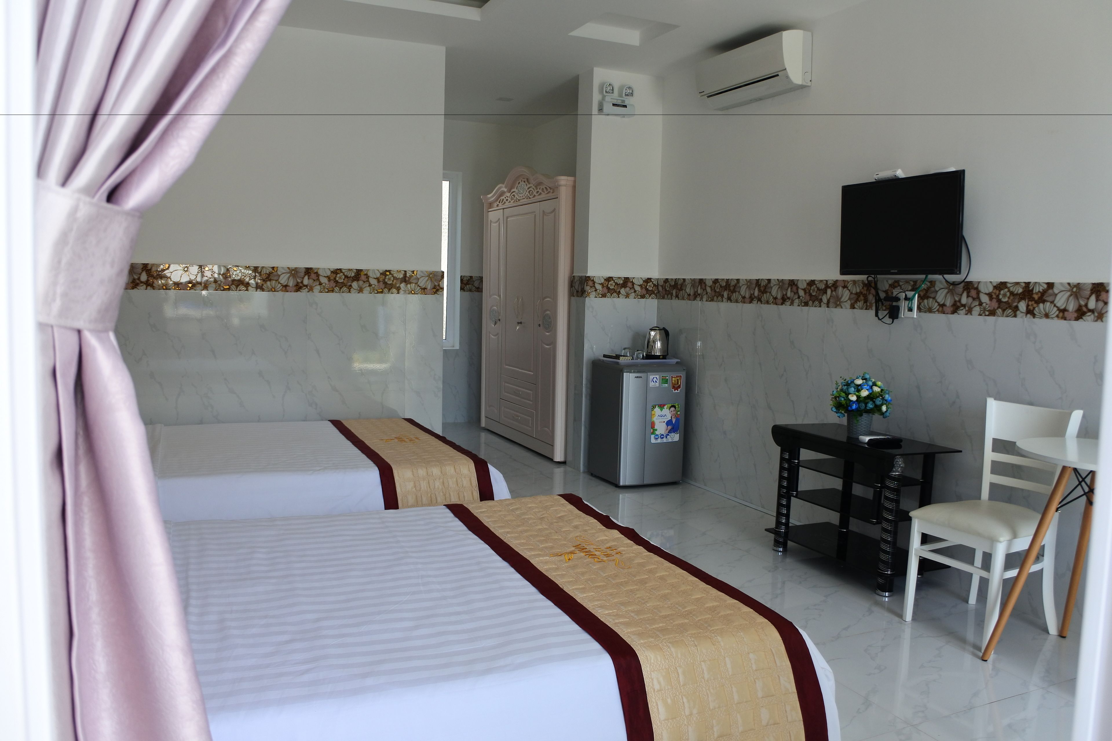 A hotel room with two beds, a TV, a refrigerator, a small table with a flower vase, and a chair.