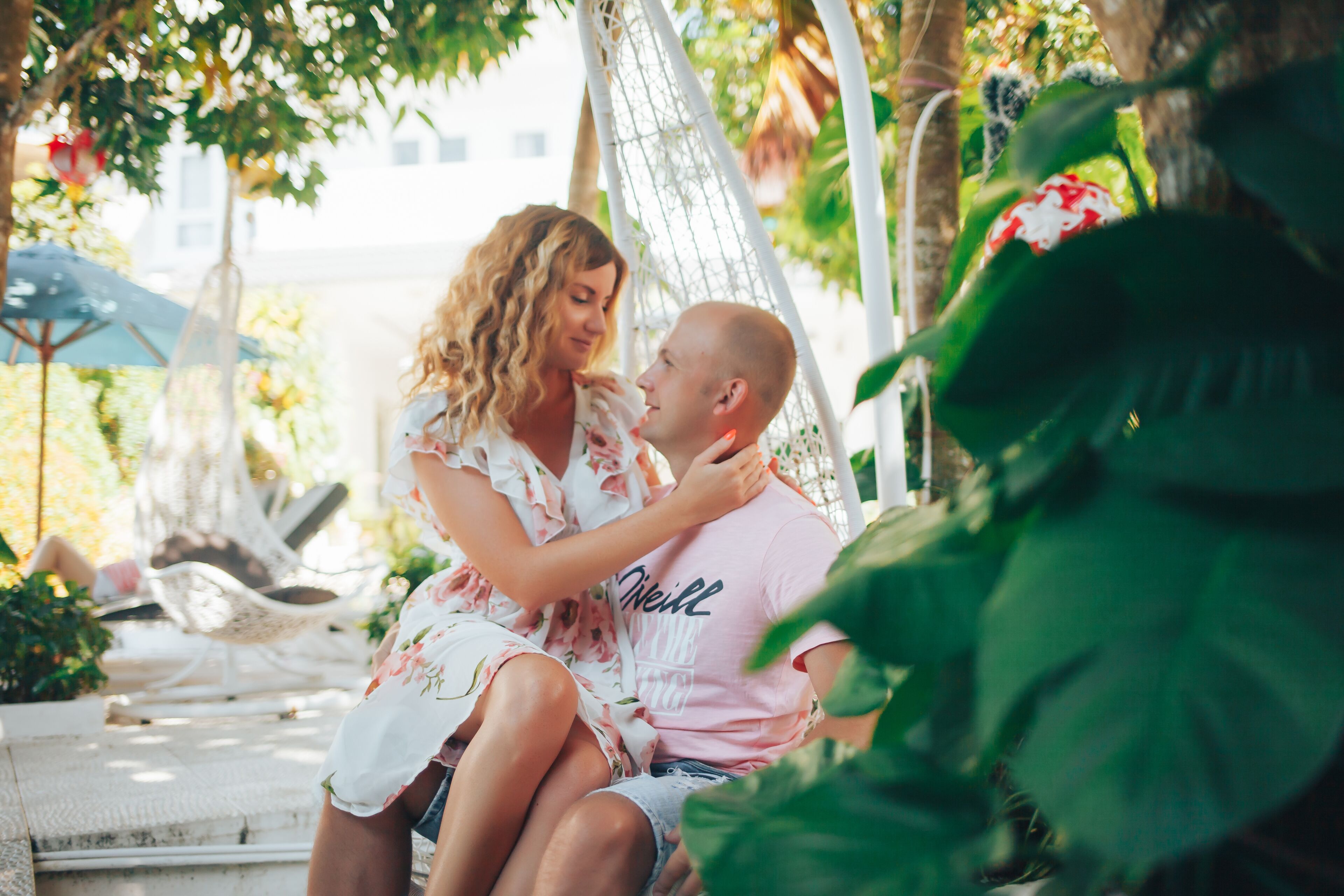A couple sitting on a hammock in a lush, green outdoor area with a white chair and umbrella in the background.