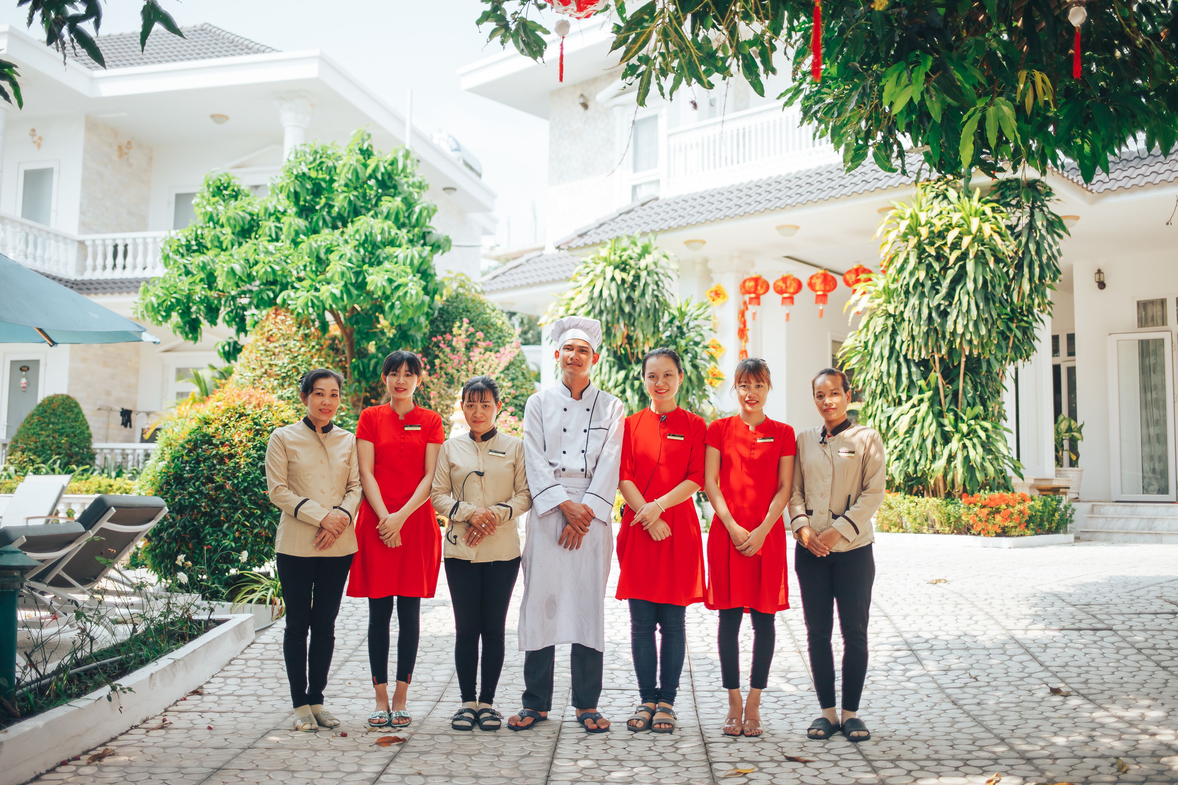 A group of people standing in a courtyard with a tiled floor, surrounded by greenery and white buildings.