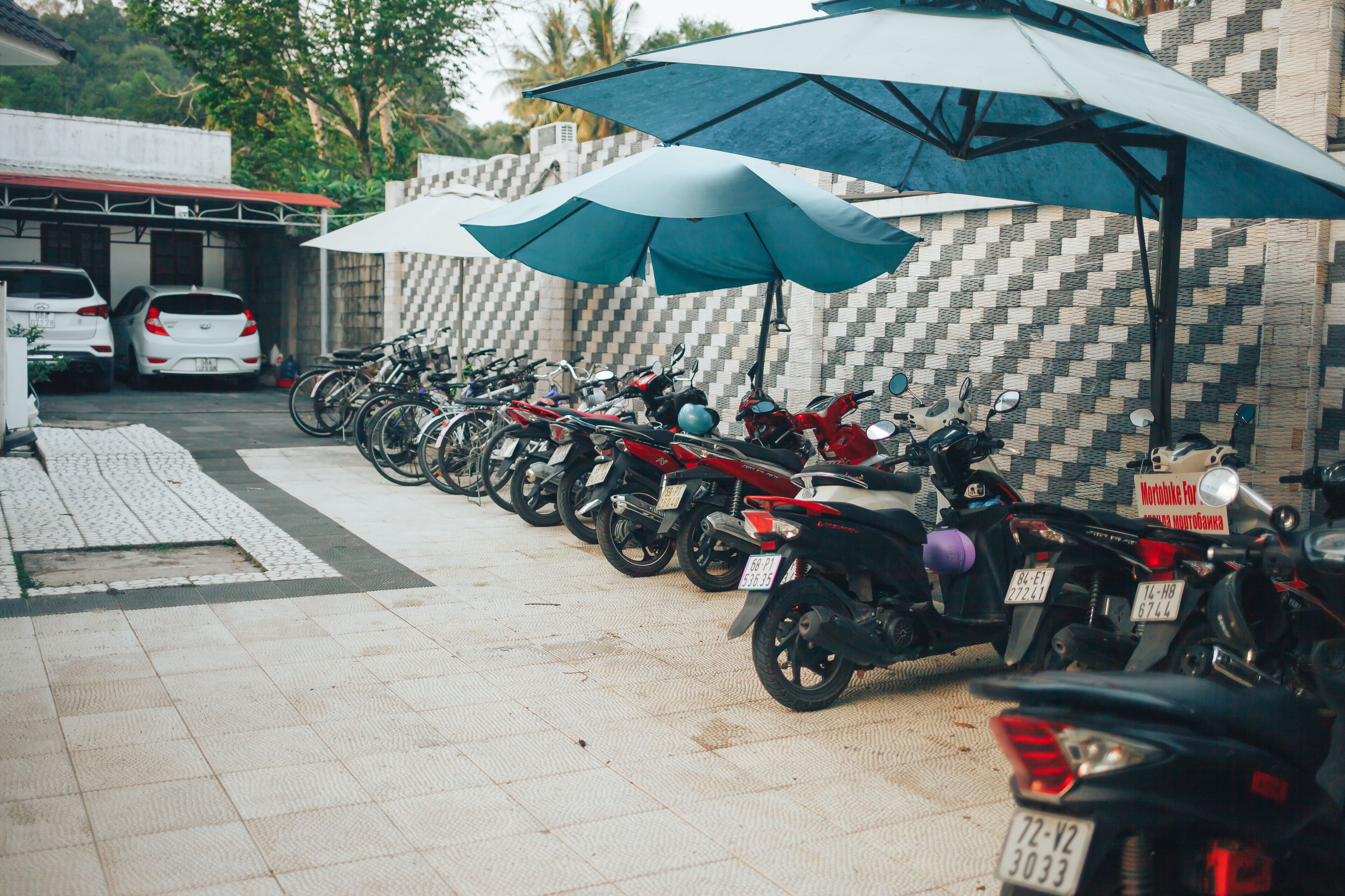 Une rangée de motos garées sous des parasols, une voiture blanche et, au fond, un bâtiment dont la façade est ornée d’un motif à carreaux.