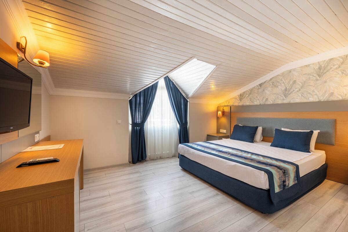 A modern hotel room with a large bed, a desk, a TV, and a skylight.