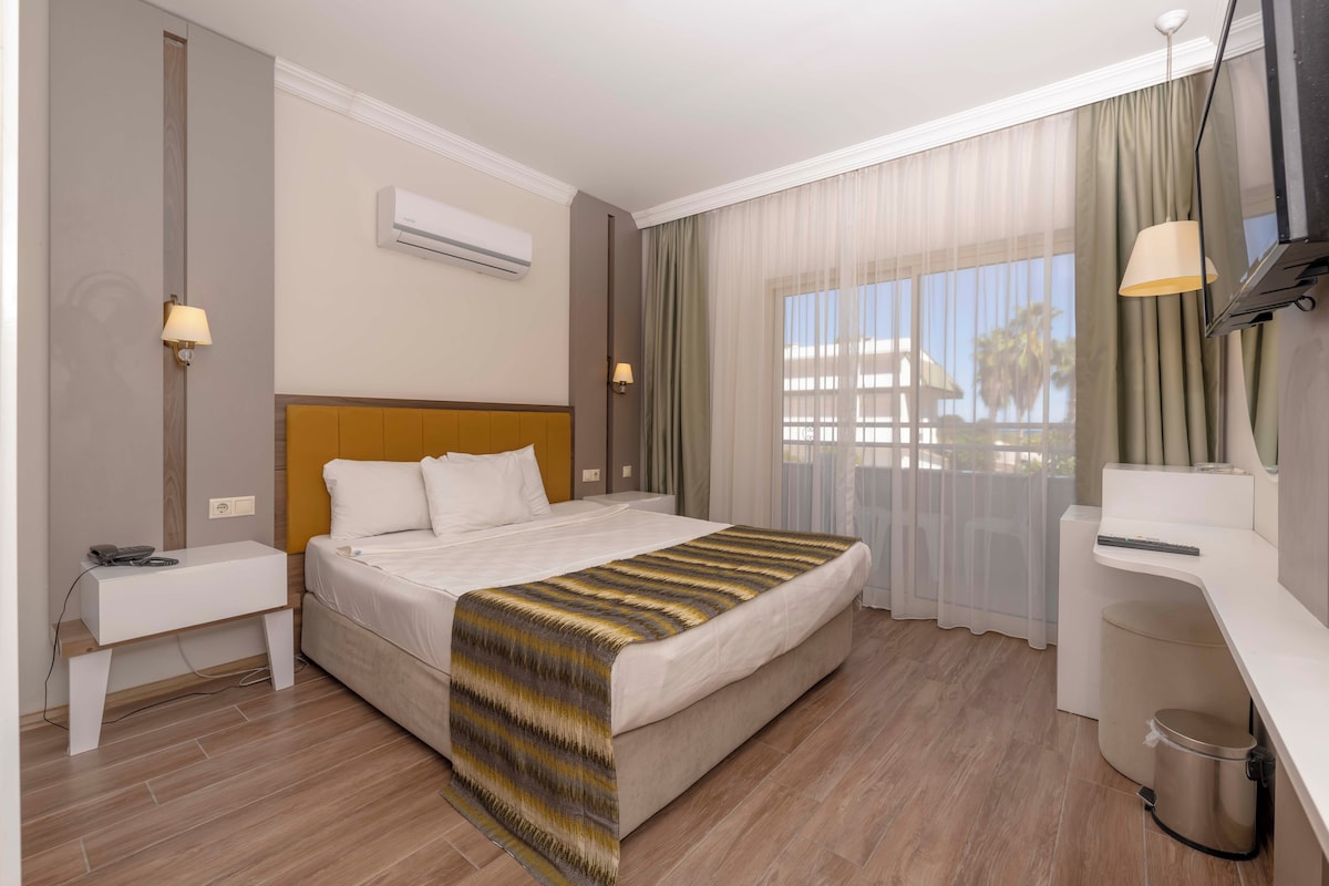 A hotel room with a bed, bedside table, lamp, air conditioning unit, and a view of the outdoors.