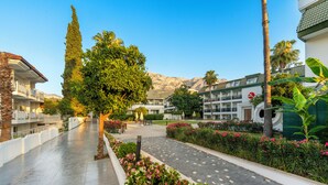 A hotel courtyard with a paved walkway, palm trees, and a mountain backdrop. - Armas Gul Beach - All Inclusive (Kemer)