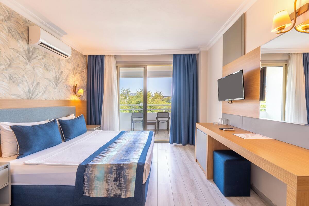 A hotel room with a large bed, a desk, a TV, and a balcony with curtains.