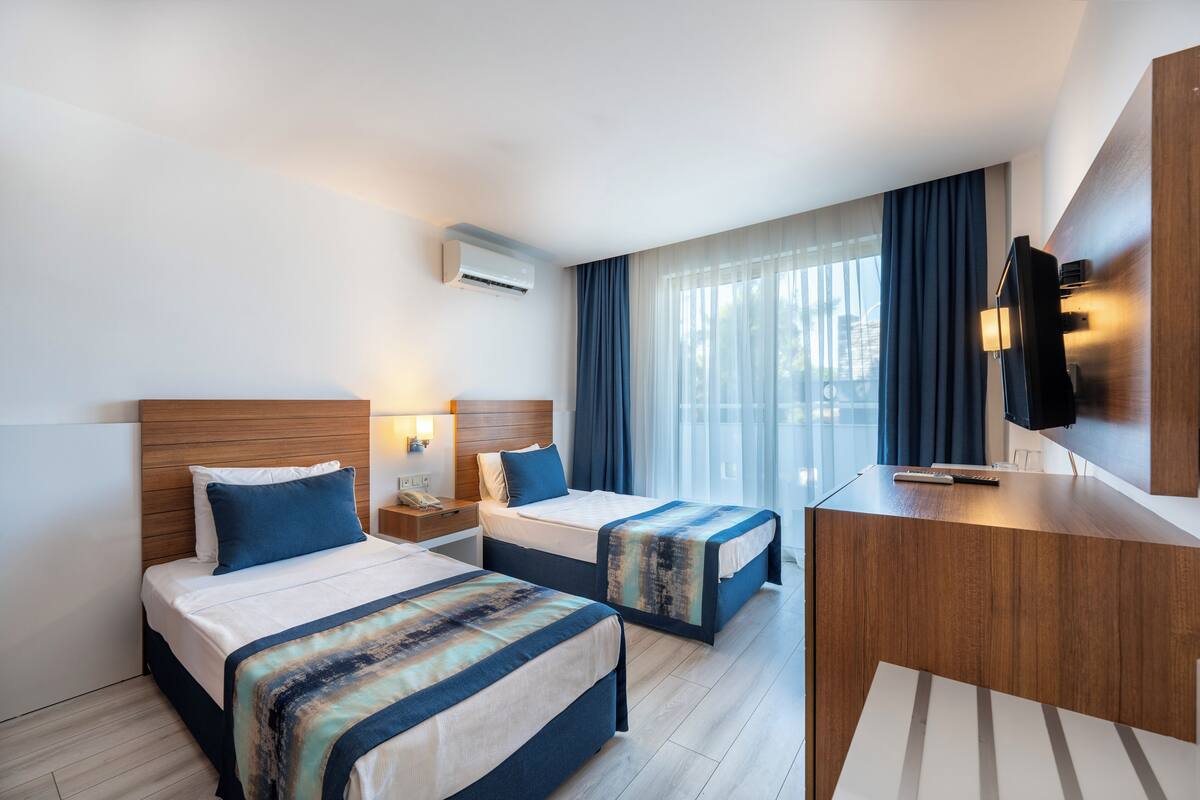 A hotel room with two beds, a desk, a TV, and a window with curtains.