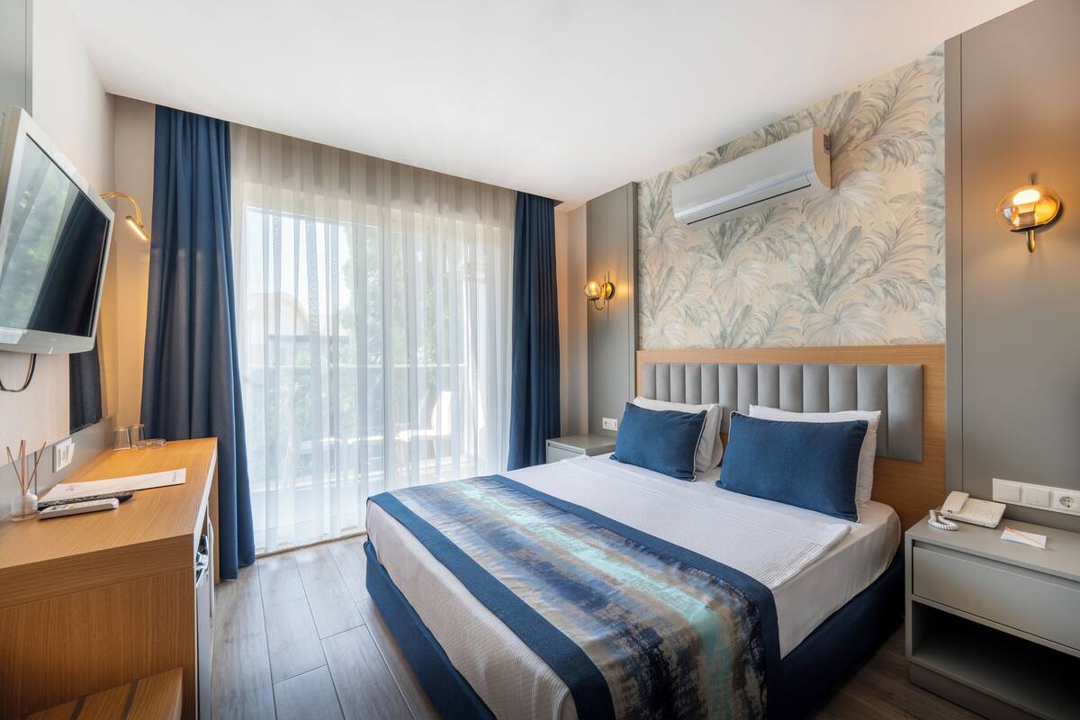 A modern hotel room with a large bed, a desk, a television, and a window with curtains.